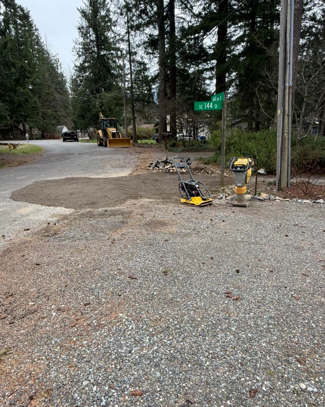 A gravel construction site with a yellow tamper and a backhoe parked in the background near a street sign.