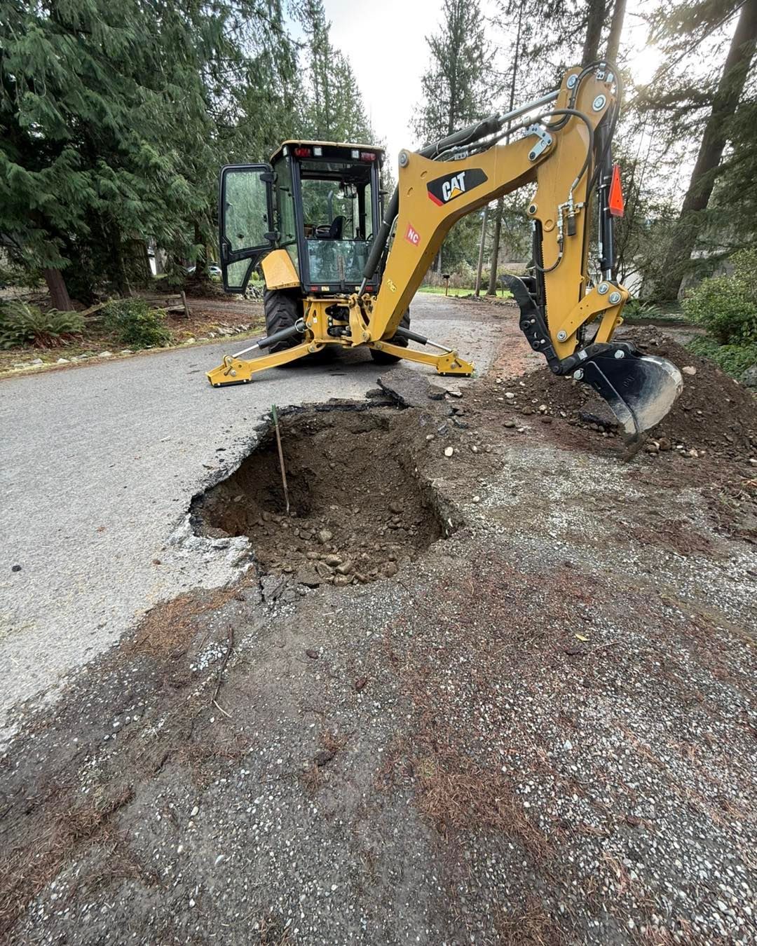 Yellow CAT backhoe parked on a gravel road next to a freshly excavated hole in the ground.
