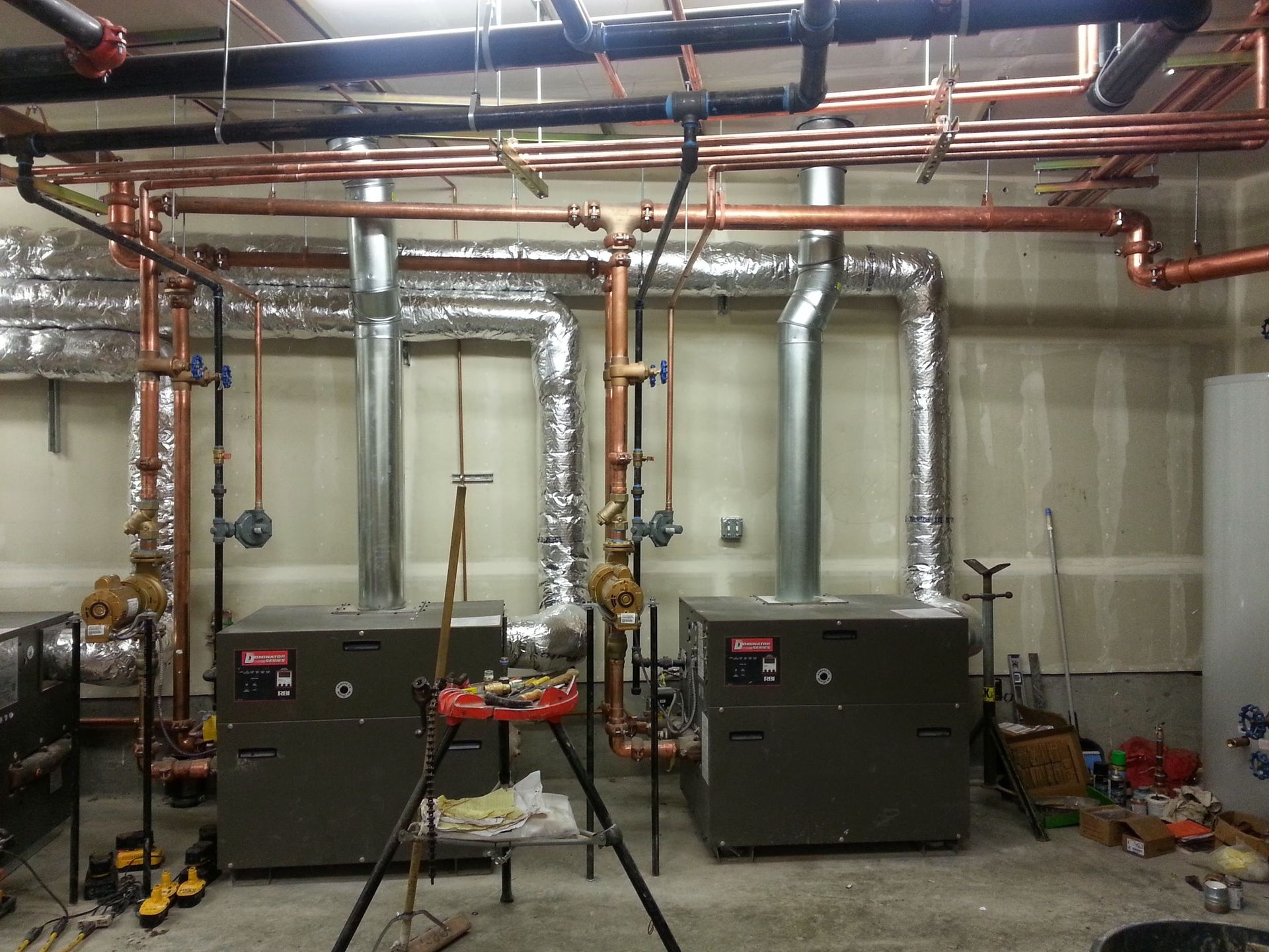 Two industrial boilers with metal exhaust vents sit in a utility room filled with copper piping and silver insulation.