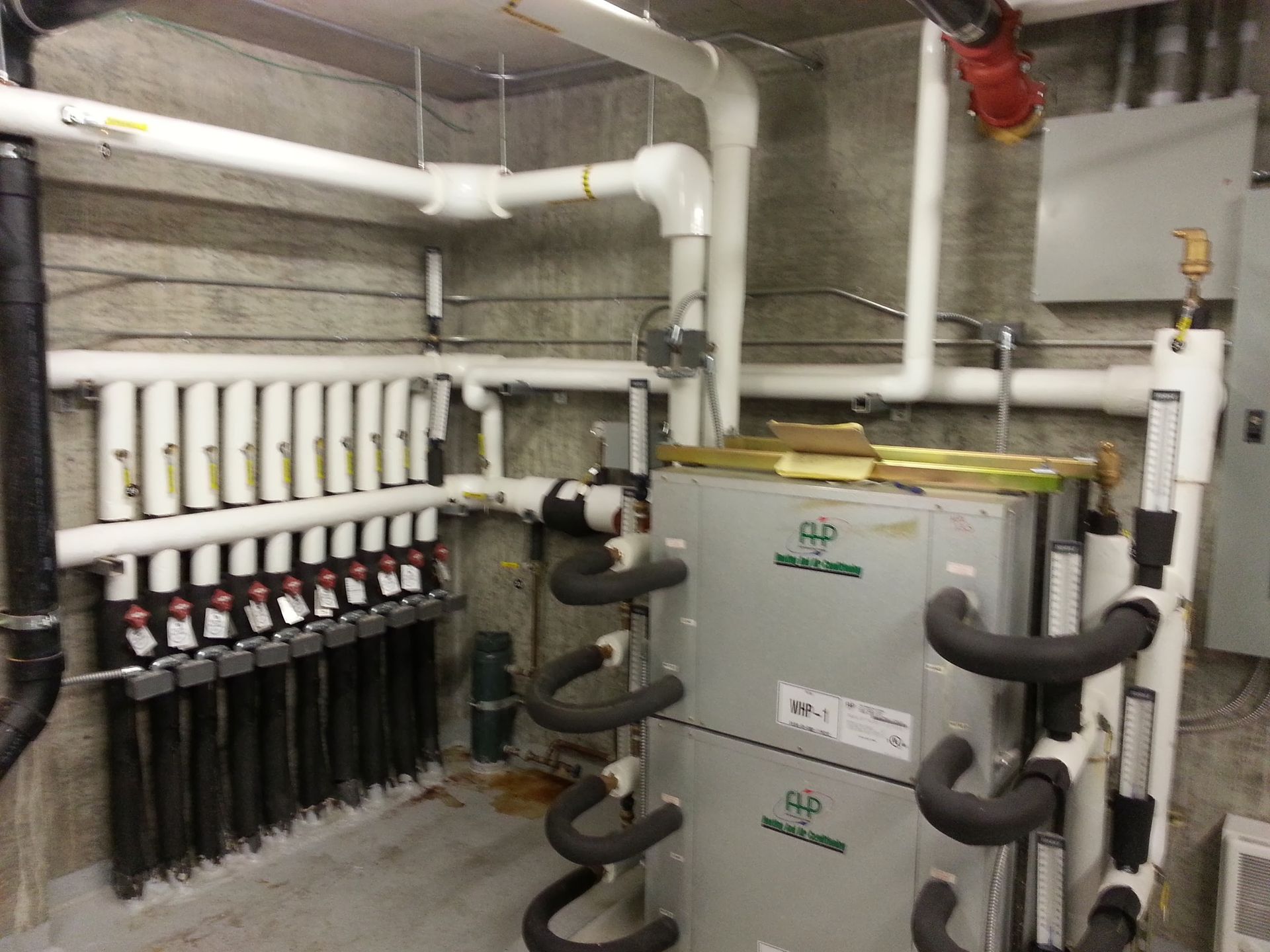 A mechanical room featuring a multi-zone radiant heating manifold with white piping and two stacked grey heating units.