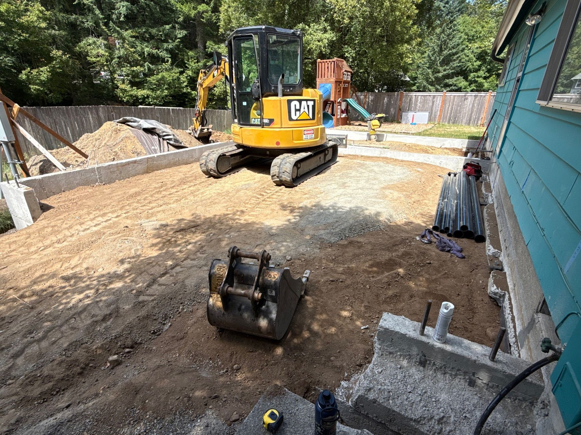 A yellow CAT mini excavator sits on a dirt construction site next to a blue-sided house with a concrete foundation.