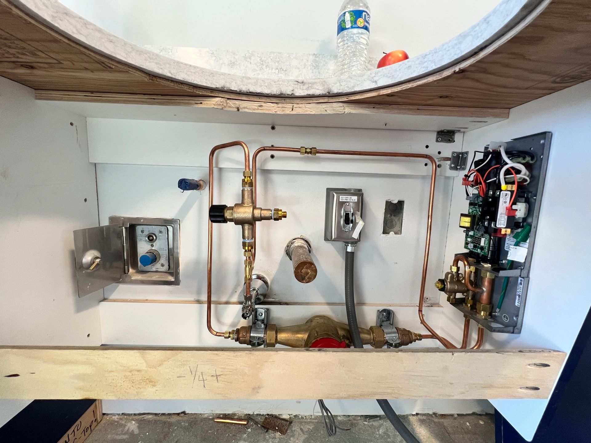 Exposed plumbing, valves, and electrical wiring inside a cabinet beneath a sink, with a water bottle resting on top.