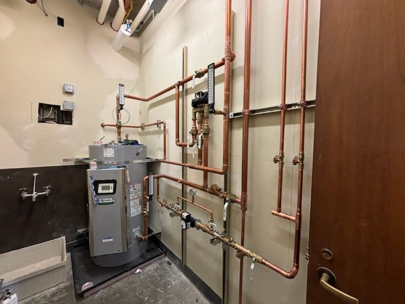A commercial water heater connected to a network of copper plumbing pipes against a utility room wall.