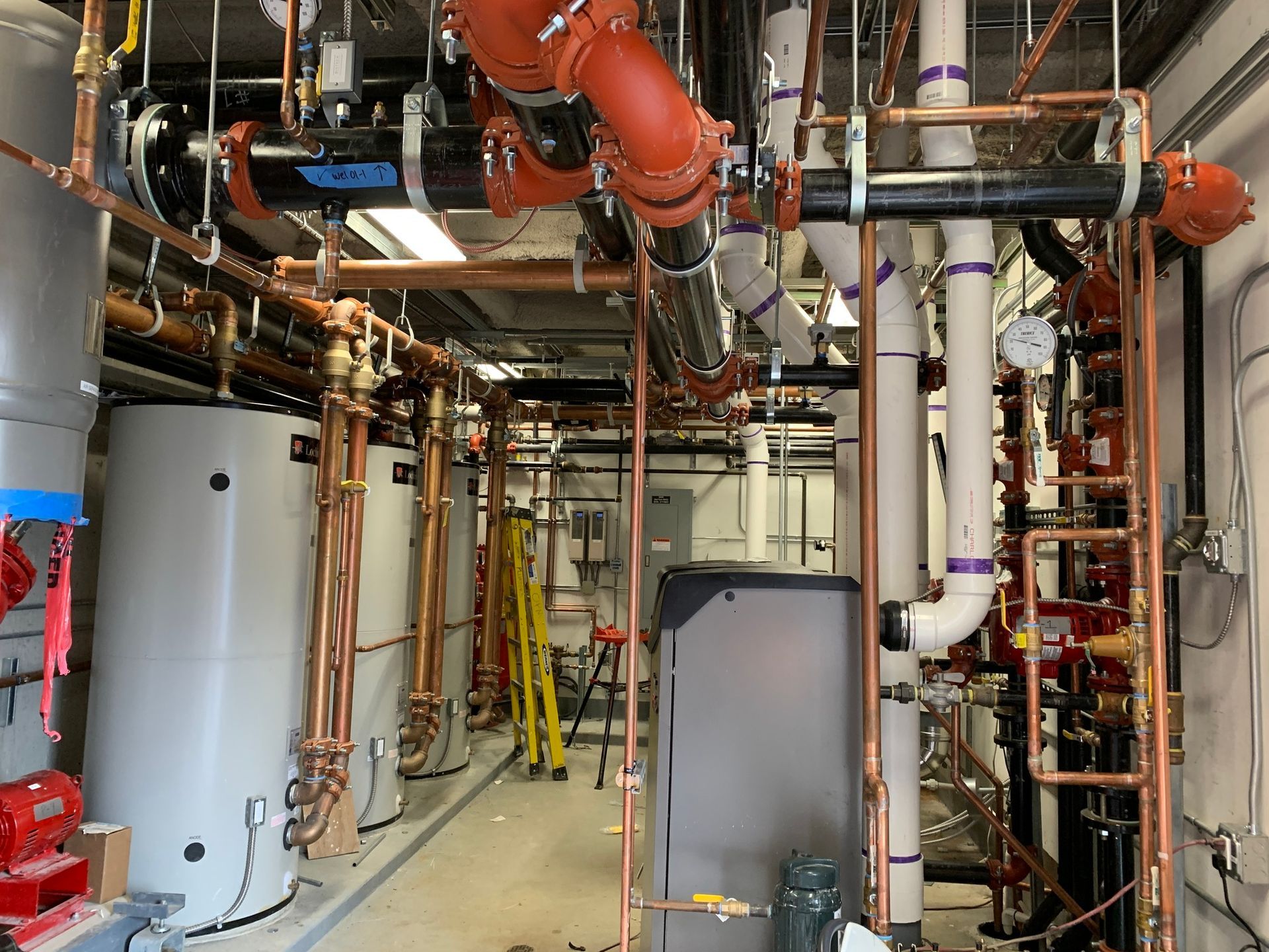 Mechanical room with white water heaters, copper piping, and black overhead conduits against a white ceiling.