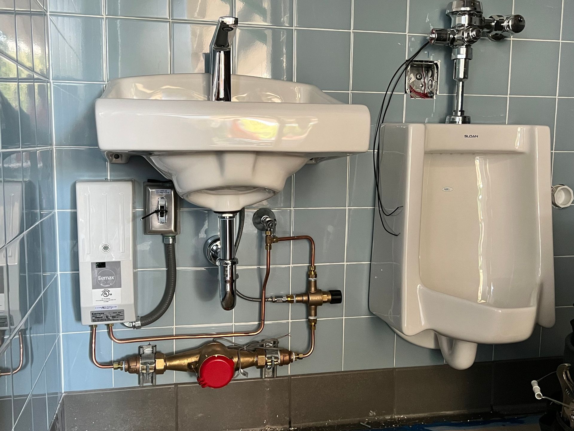 A white sink and urinal mounted on a light blue tiled wall, with exposed copper plumbing and electrical components.