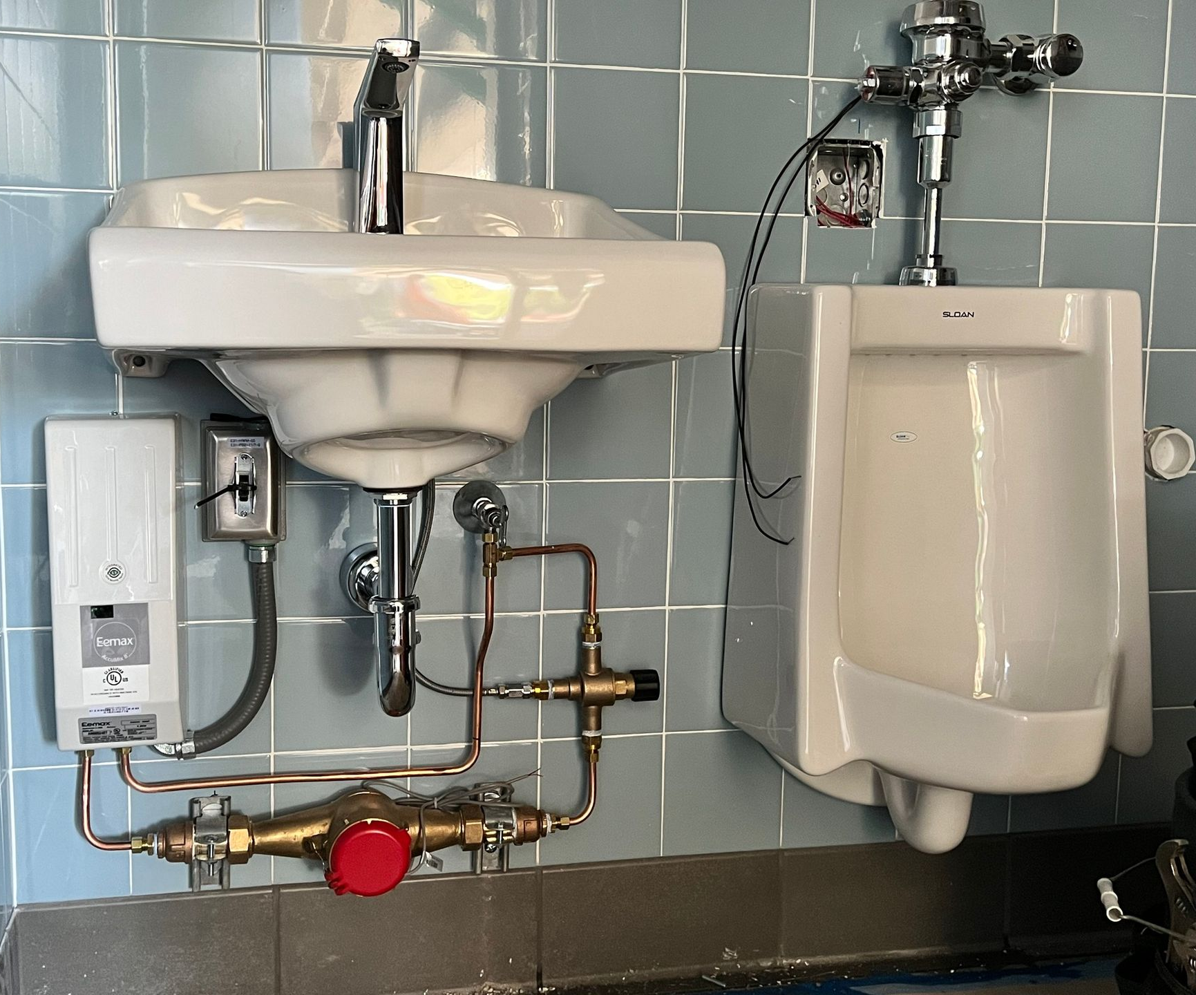 A wall-mounted sink with exposed plumbing next to a urinal against a blue-tiled wall in a commercial restroom.