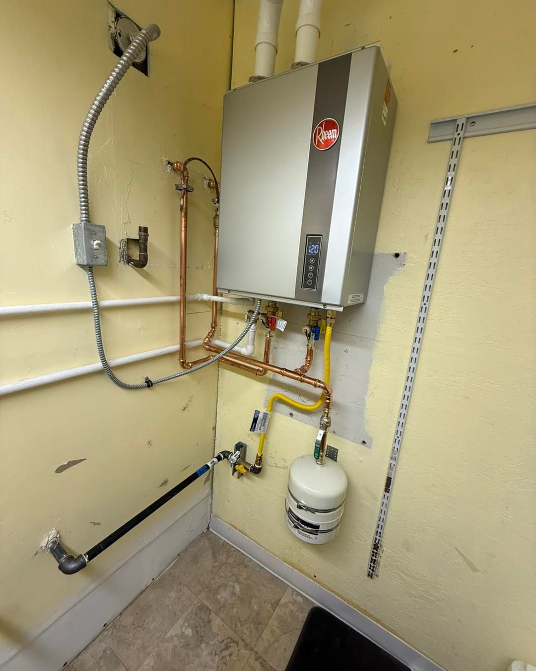A Rheem tankless water heater mounted on a yellow wall with exposed copper plumbing, a gas line, and a small pressure tank.