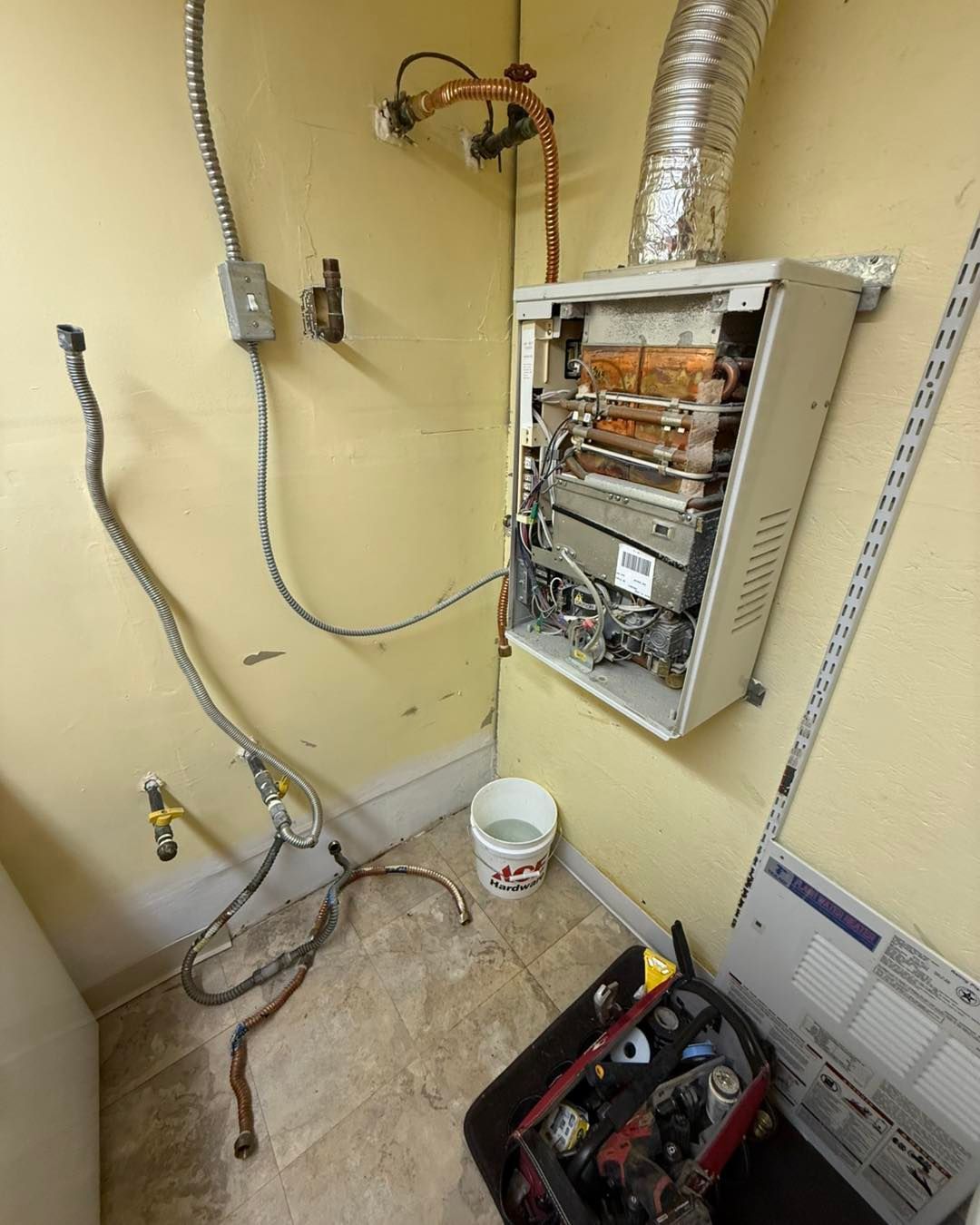 An open tankless water heater mounted on a yellow wall with exposed wiring, plumbing pipes, and a bucket on the floor.