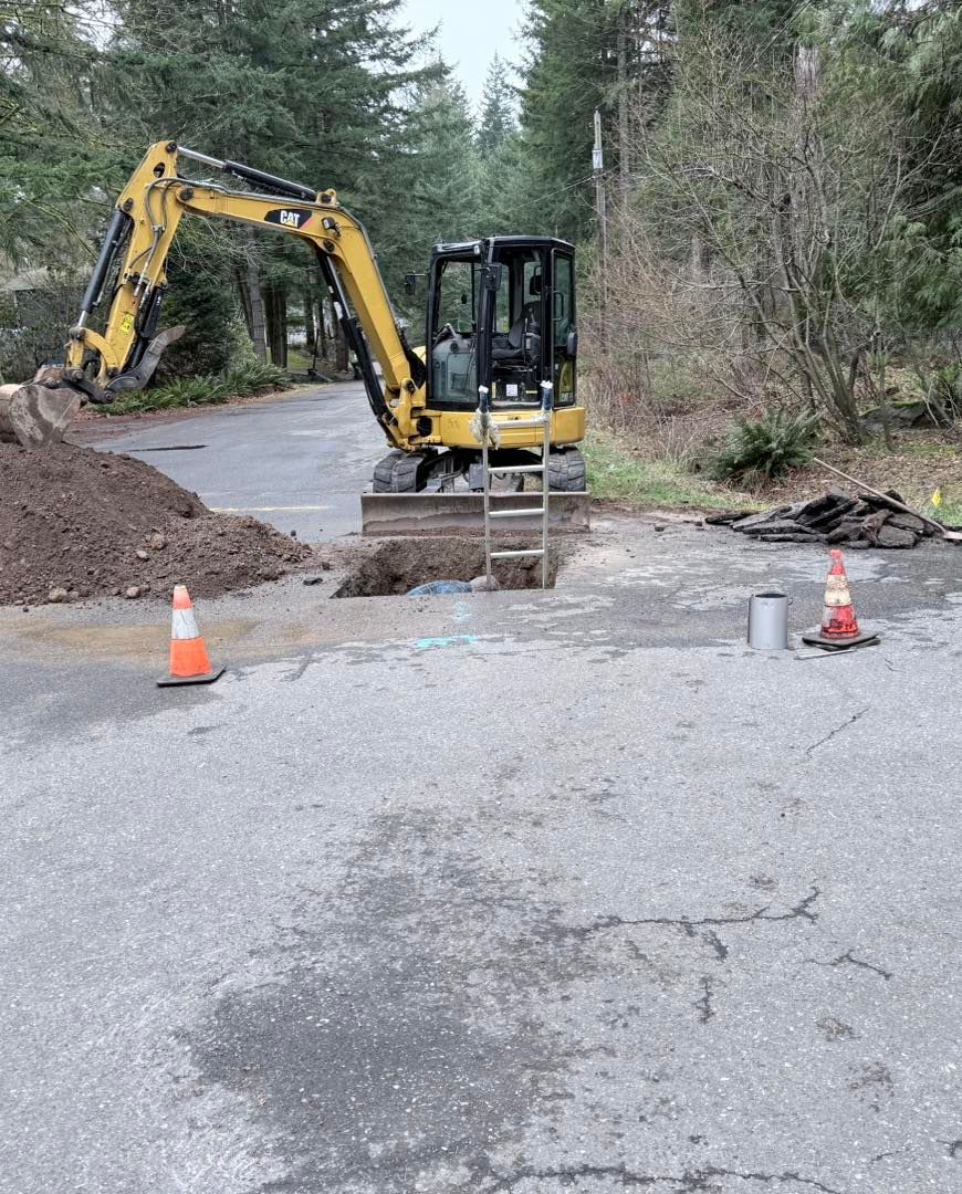 A yellow excavator parked on an asphalt road next to a trench with a ladder, marked by two orange safety cones.