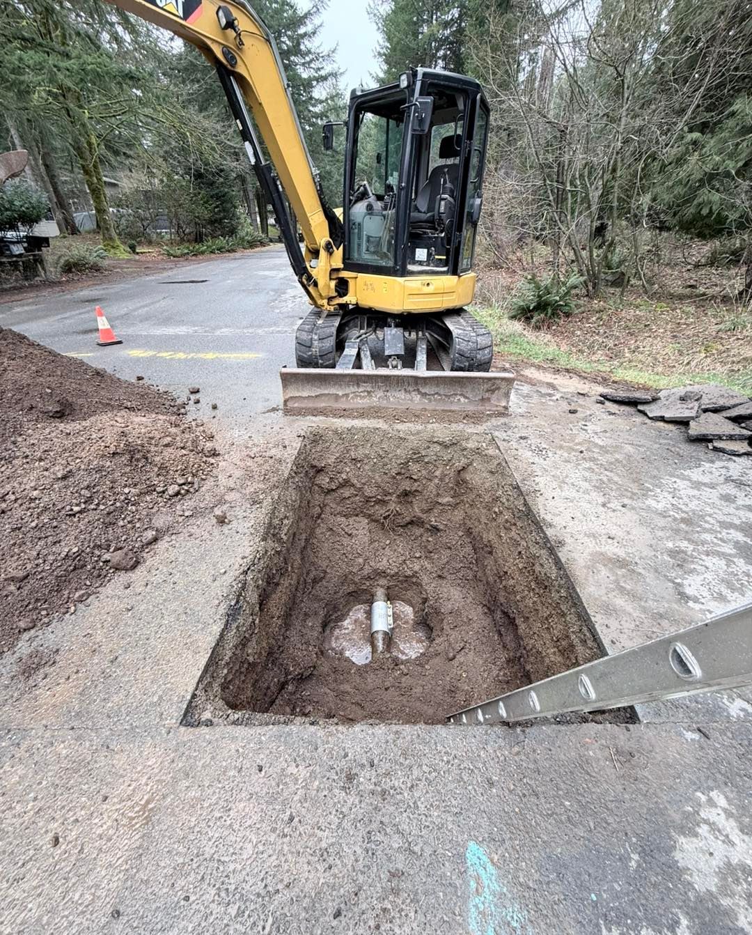 A yellow excavator parked behind a rectangular excavation pit in a paved area, with a ladder leaning into the hole.