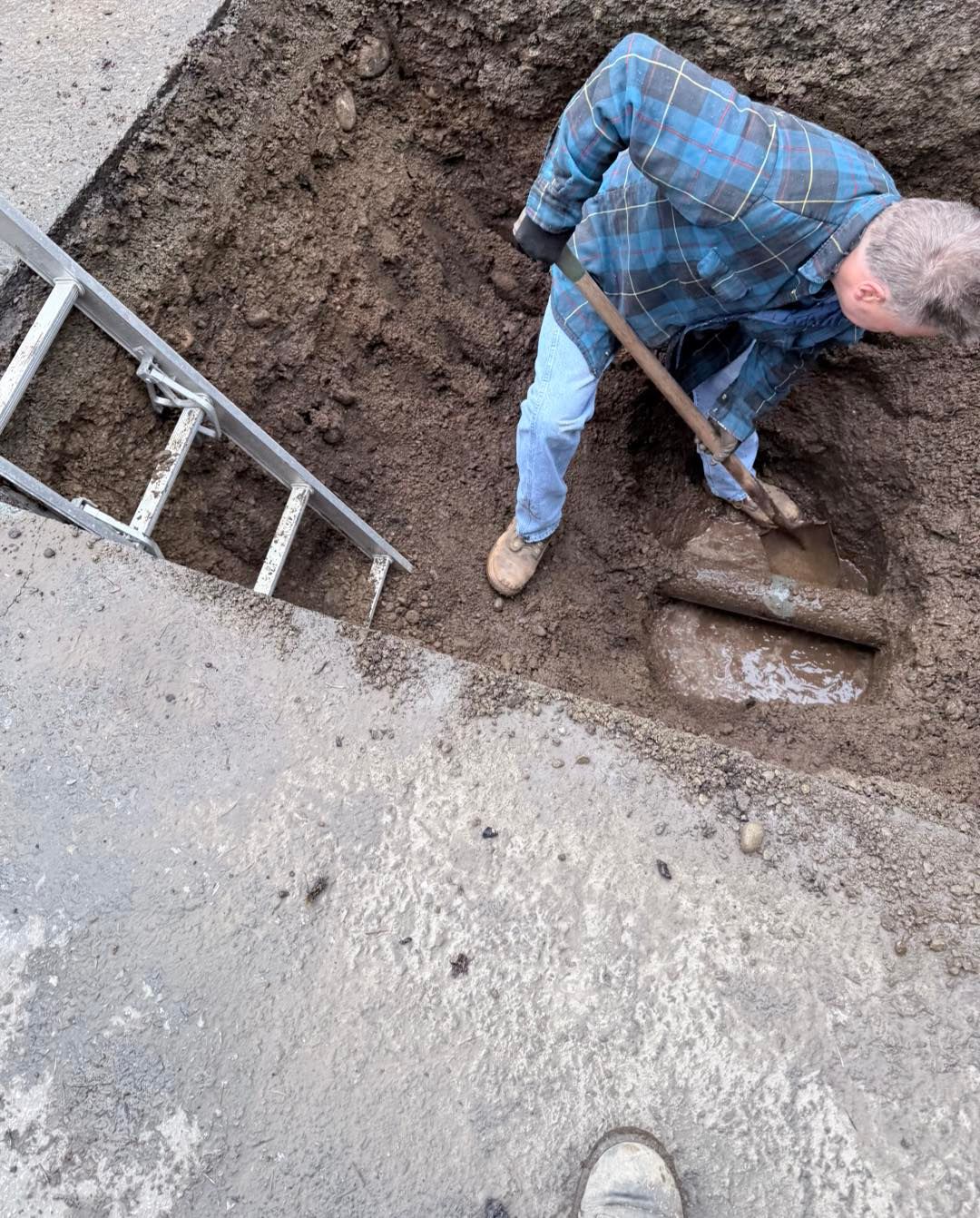 A person in a blue flannel shirt uses a shovel to dig around a pipe in a dirt-filled trench next to a ladder.