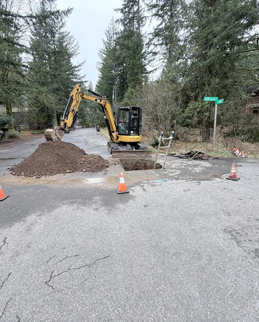 A yellow excavator parked on an asphalt road near a pile of dirt and an open trench with orange traffic cones.