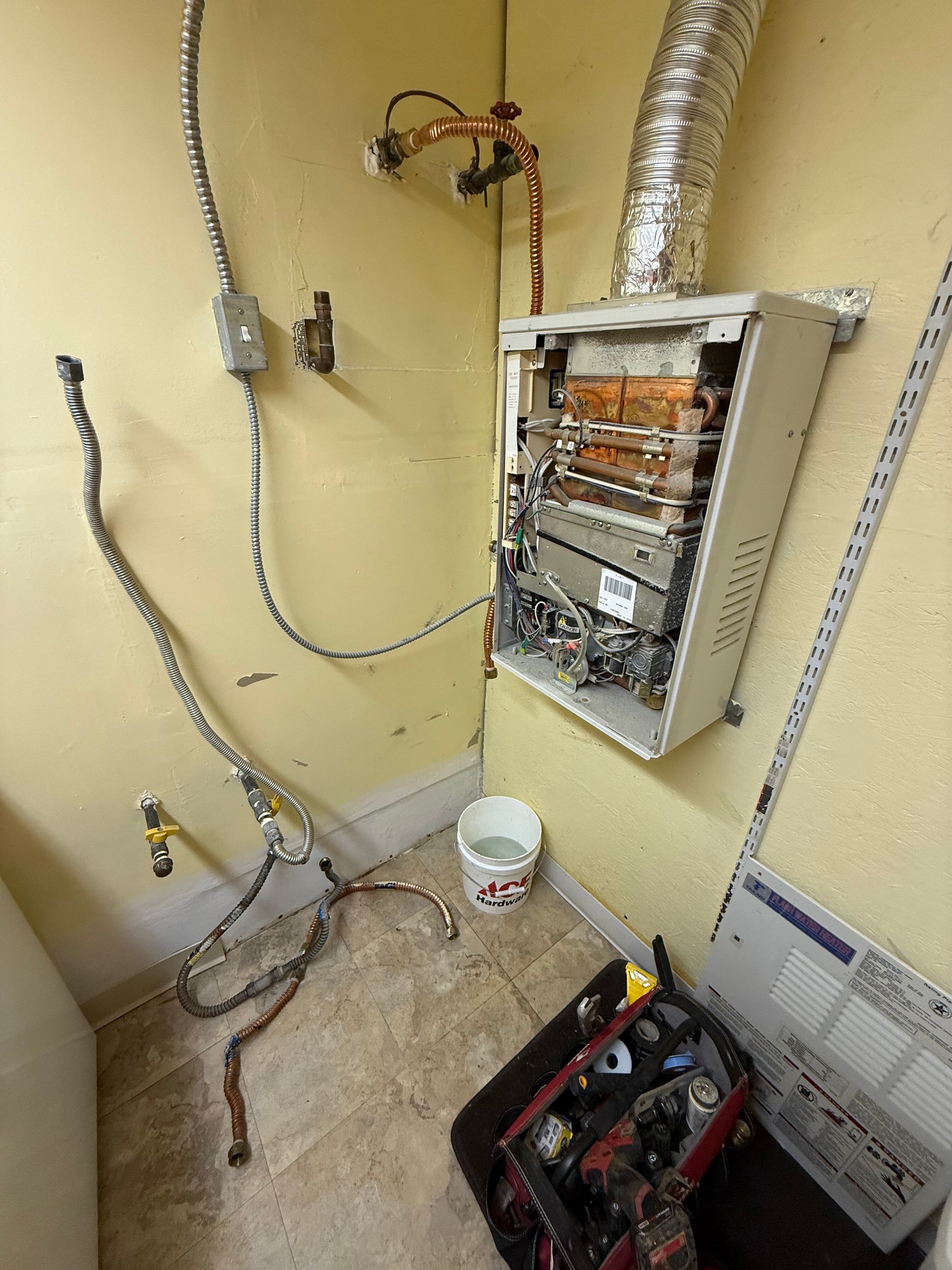 A tankless water heater with exposed components, disconnected hoses, and a tool bag on the floor in a utility room.