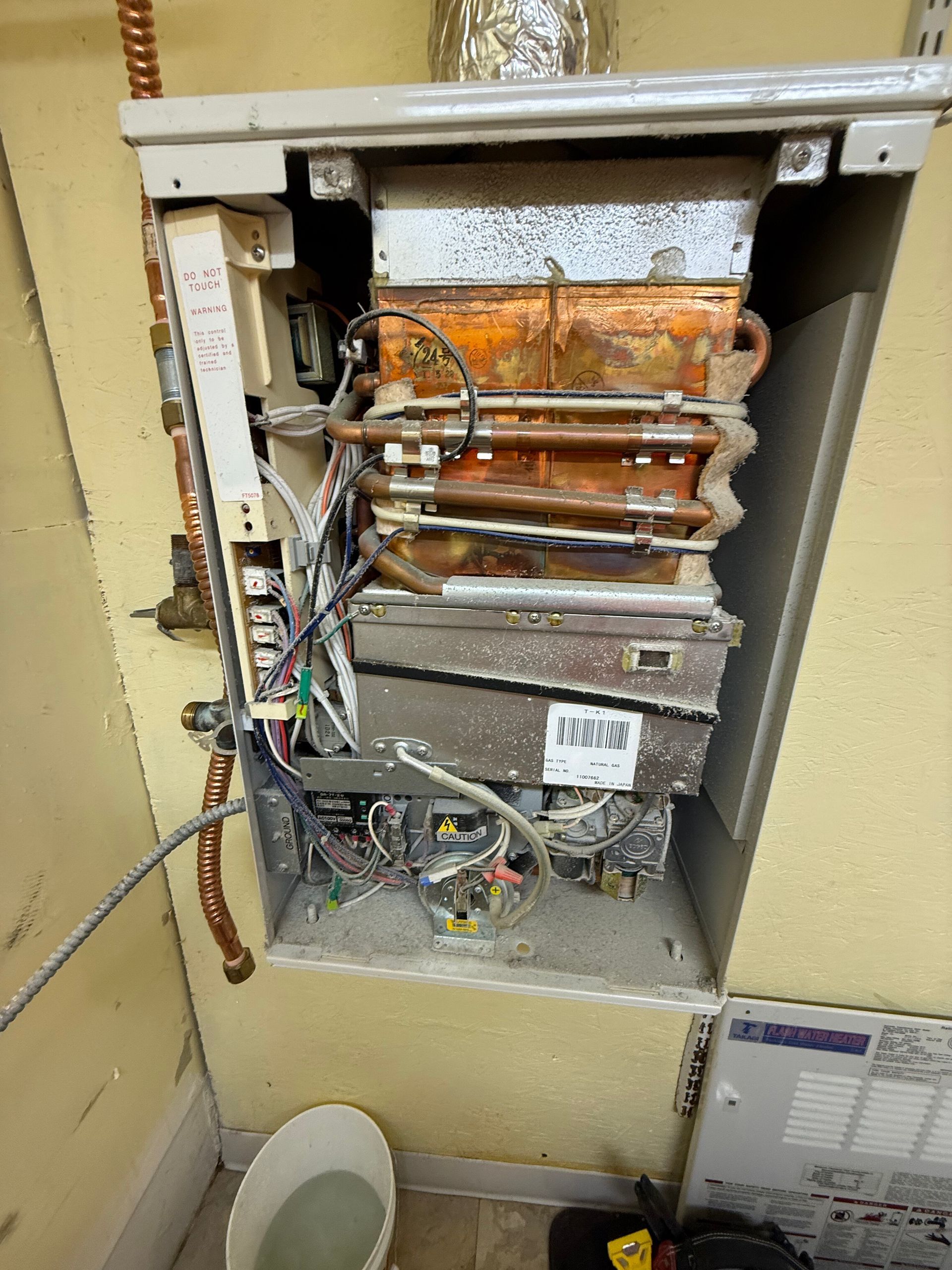 The interior components of a wall-mounted water heater with exposed copper pipes, wires, and a bucket sitting below it.