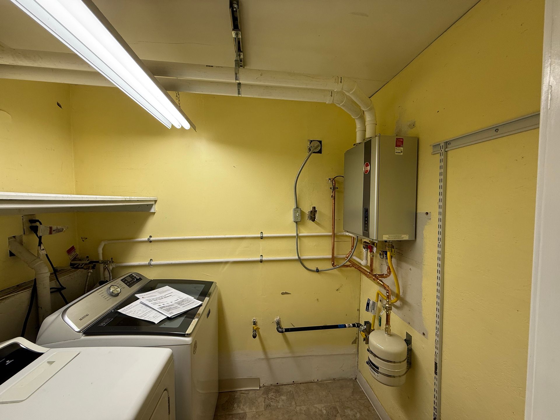 A utility room with a washing machine, a wall-mounted tankless water heater, and exposed pipes against yellow walls.