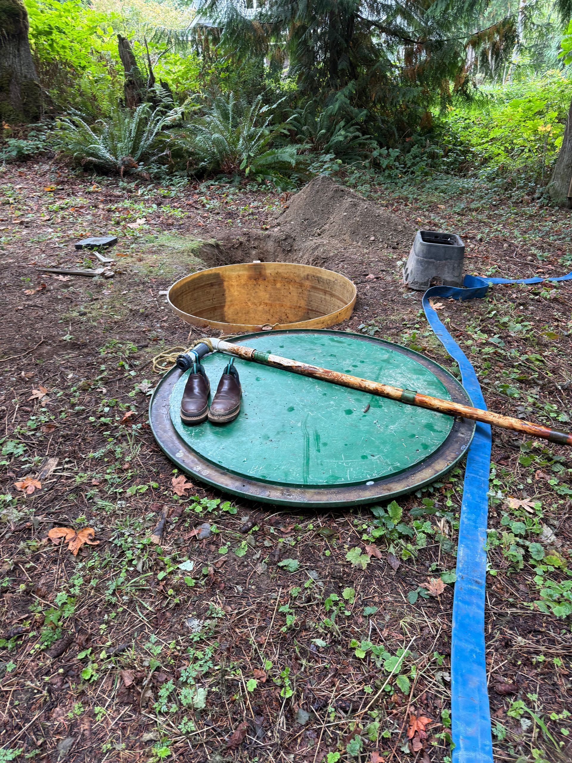 A partially opened septic tank lid sits on a forest floor with a pair of leather shoes and a long tool placed on top.