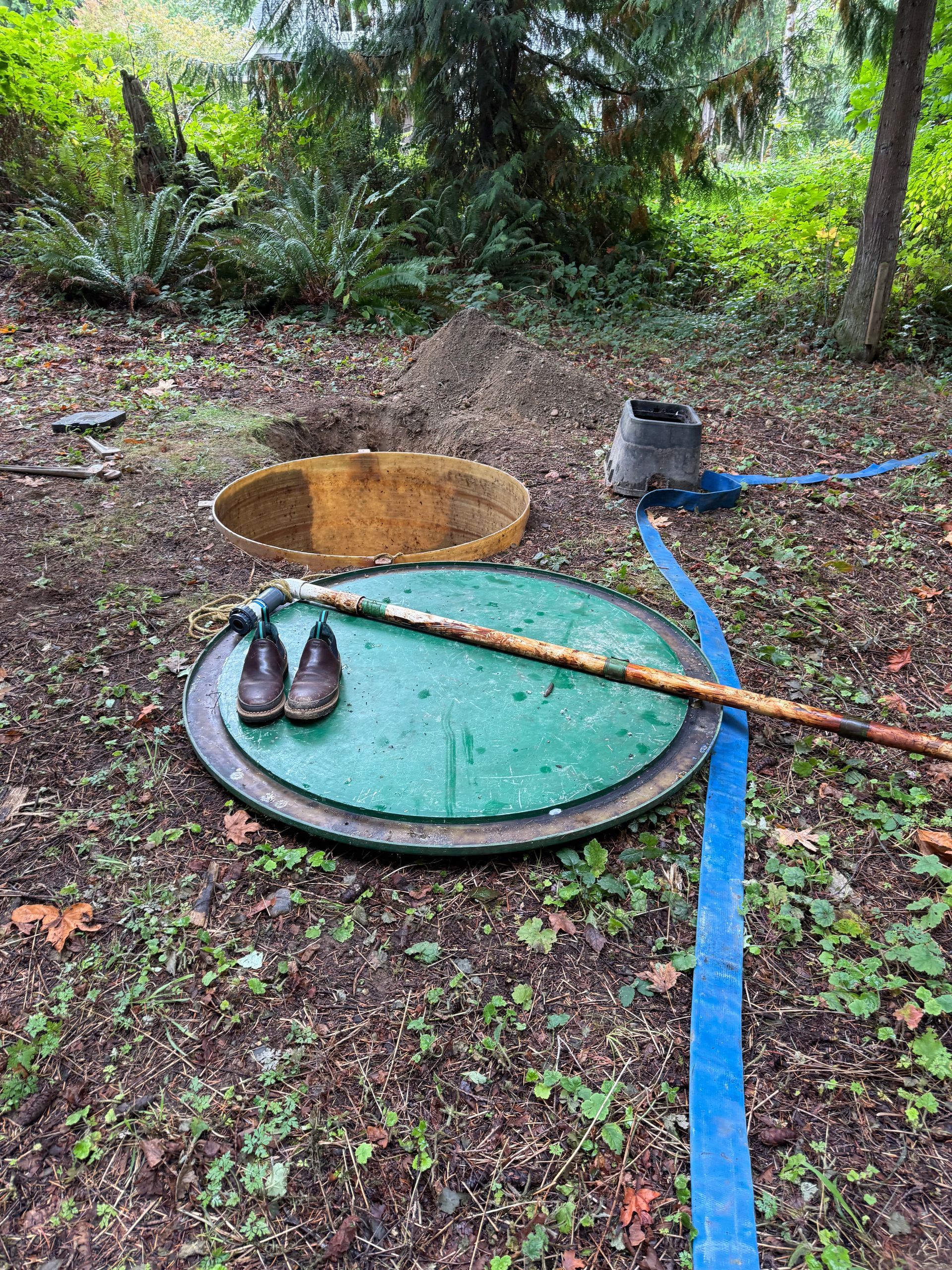 A green circular septic tank cover sits on the ground next to an open hole, a pair of boots, and a blue hose in a forest.