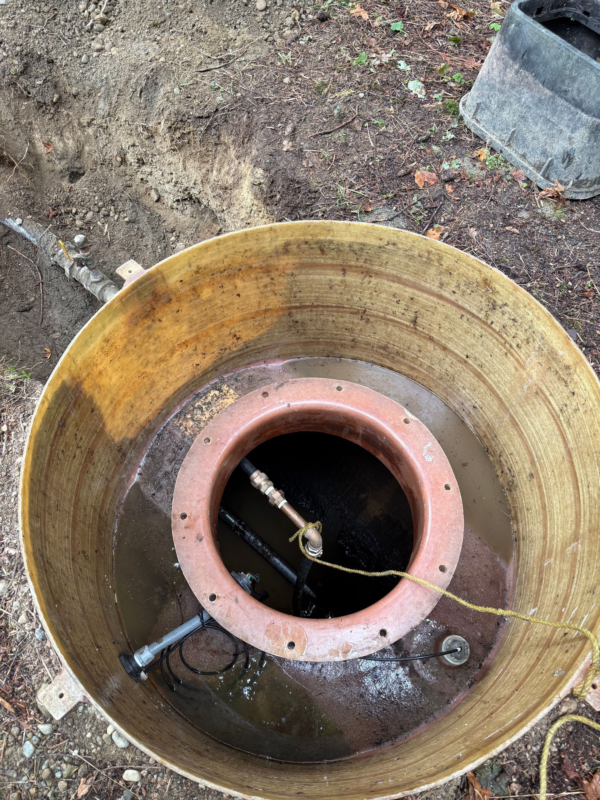 An open, round outdoor utility well filled with murky water, showing interior piping and a red cylindrical sleeve.