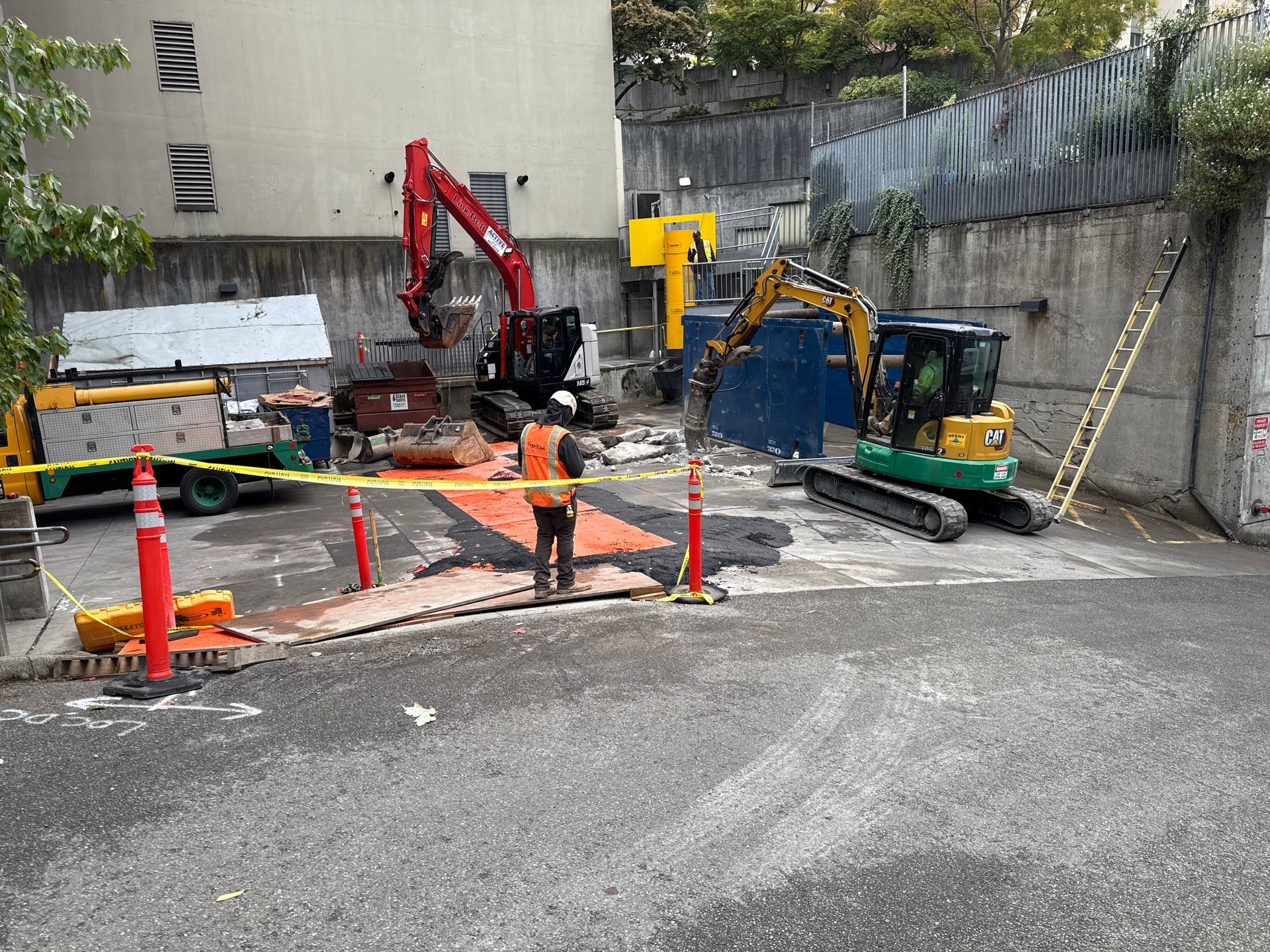 A construction site with two excavators, a worker in a safety vest, and orange caution tape in a concrete area.
