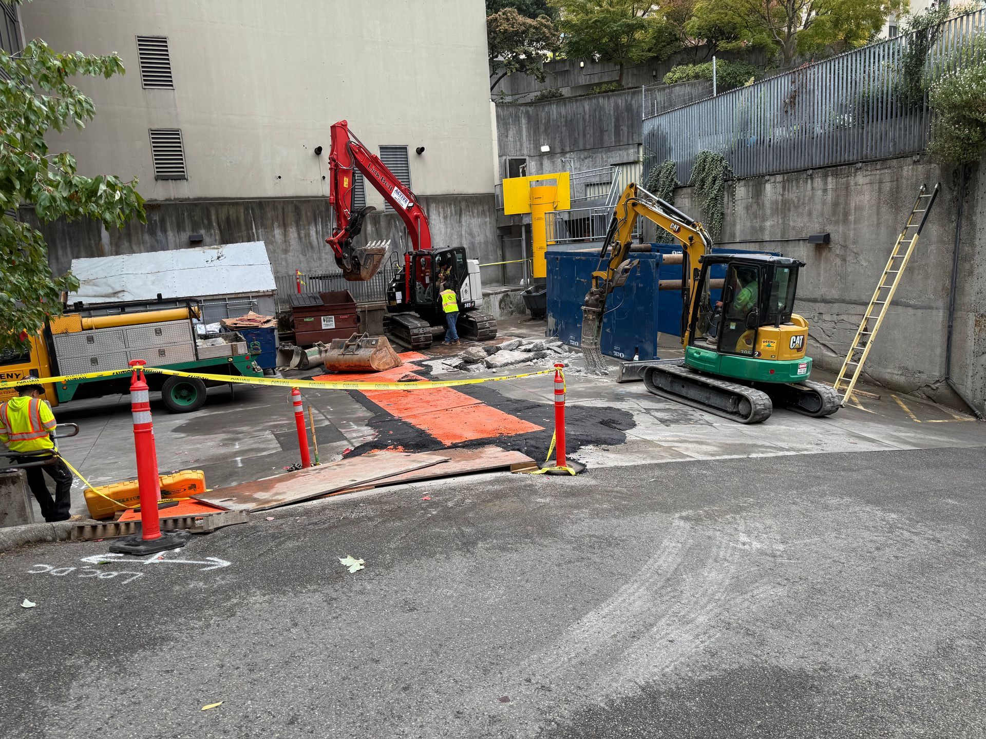Two excavators work at an urban construction site with orange warning barriers and a person in a safety vest.