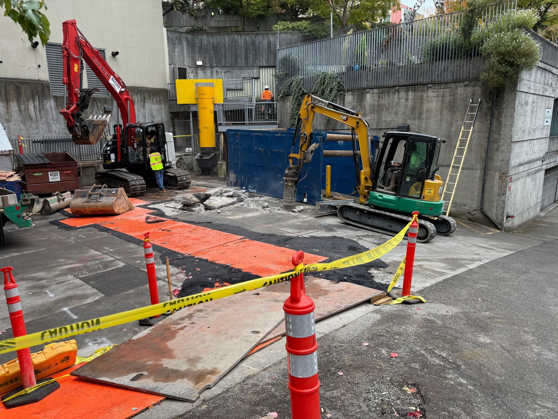 Construction site featuring red and yellow excavators, a blue dumpster, and orange safety flooring behind caution tape.