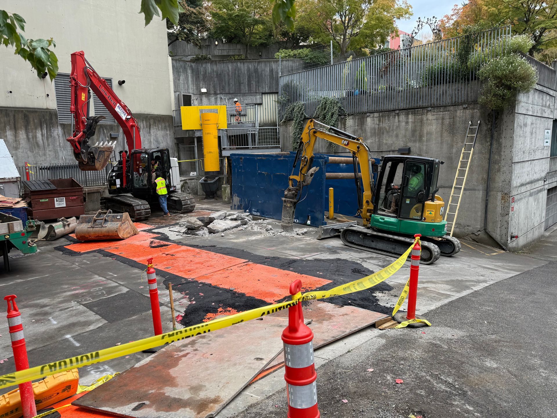 Two excavators work at an urban construction site surrounded by orange safety tape and cones.