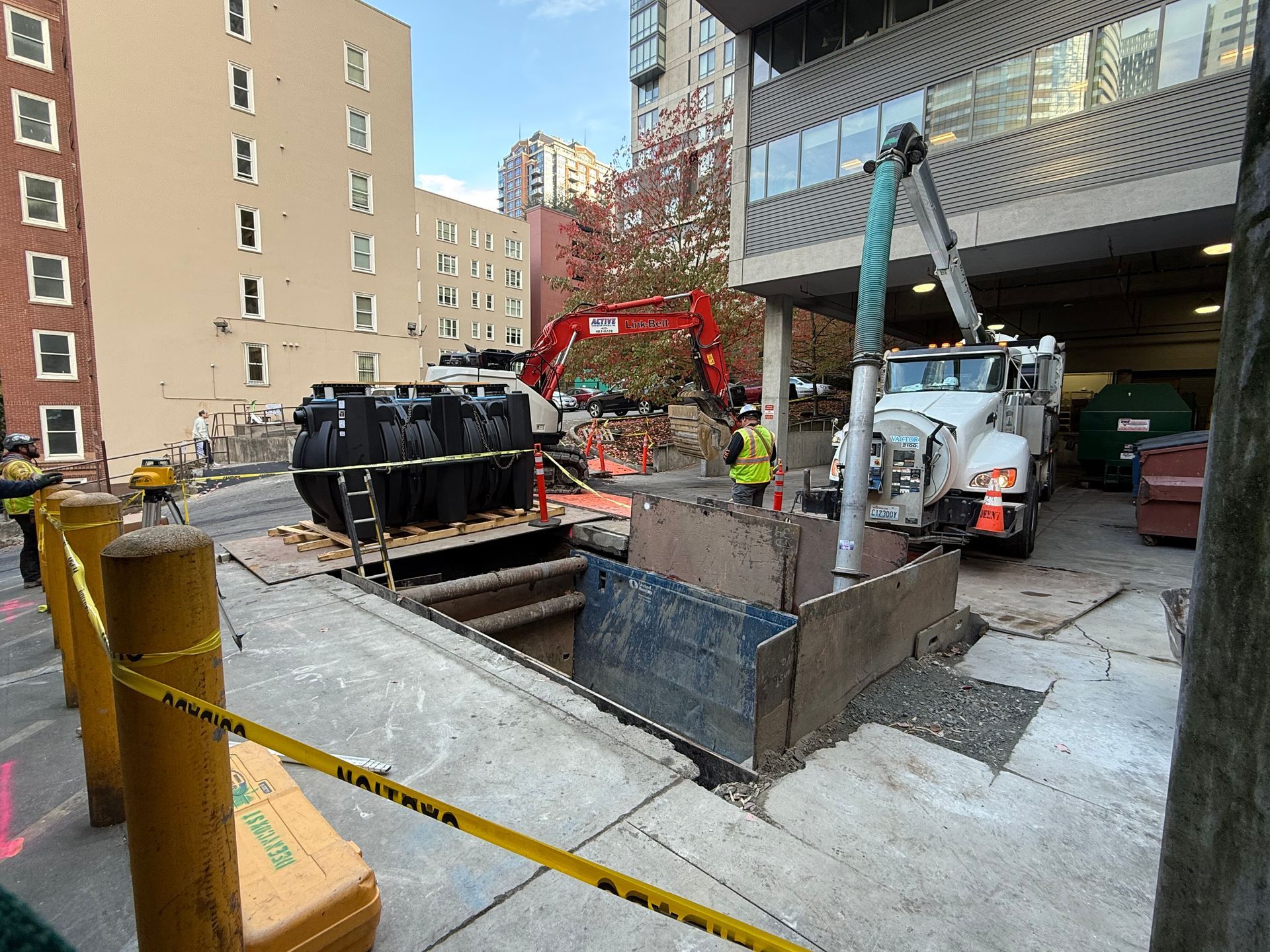 Construction site with a deep excavated trench, a white vacuum truck, and a red excavator in a city setting.
