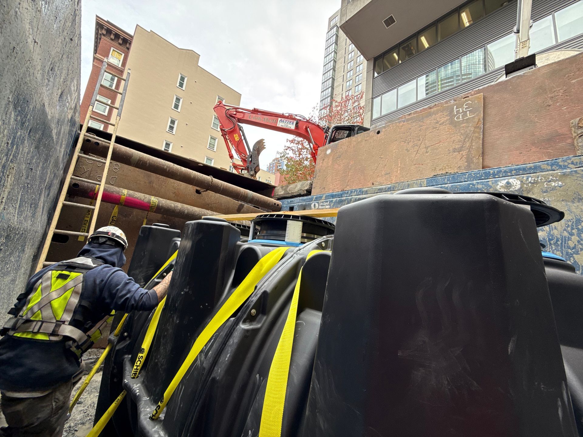 A construction worker in a high-visibility vest secures a large black tank with yellow straps at a site with an excavator.