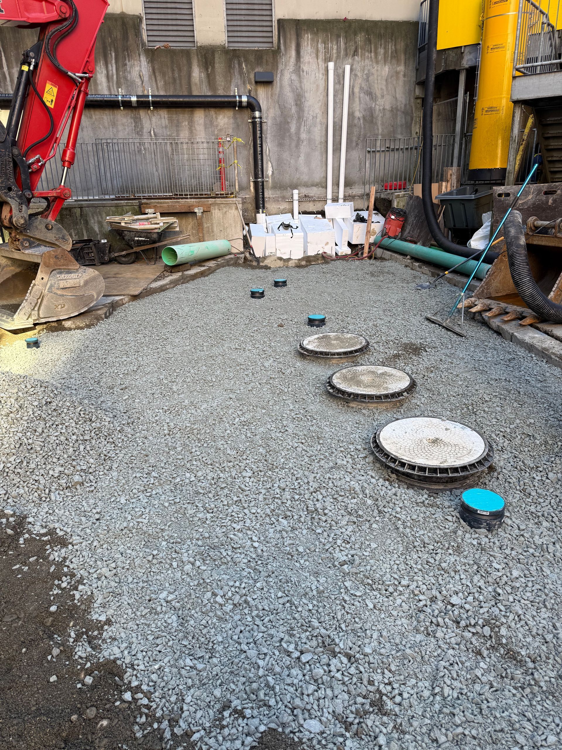 Gravel-covered construction site with several circular maintenance access covers, pipes, and heavy machinery.