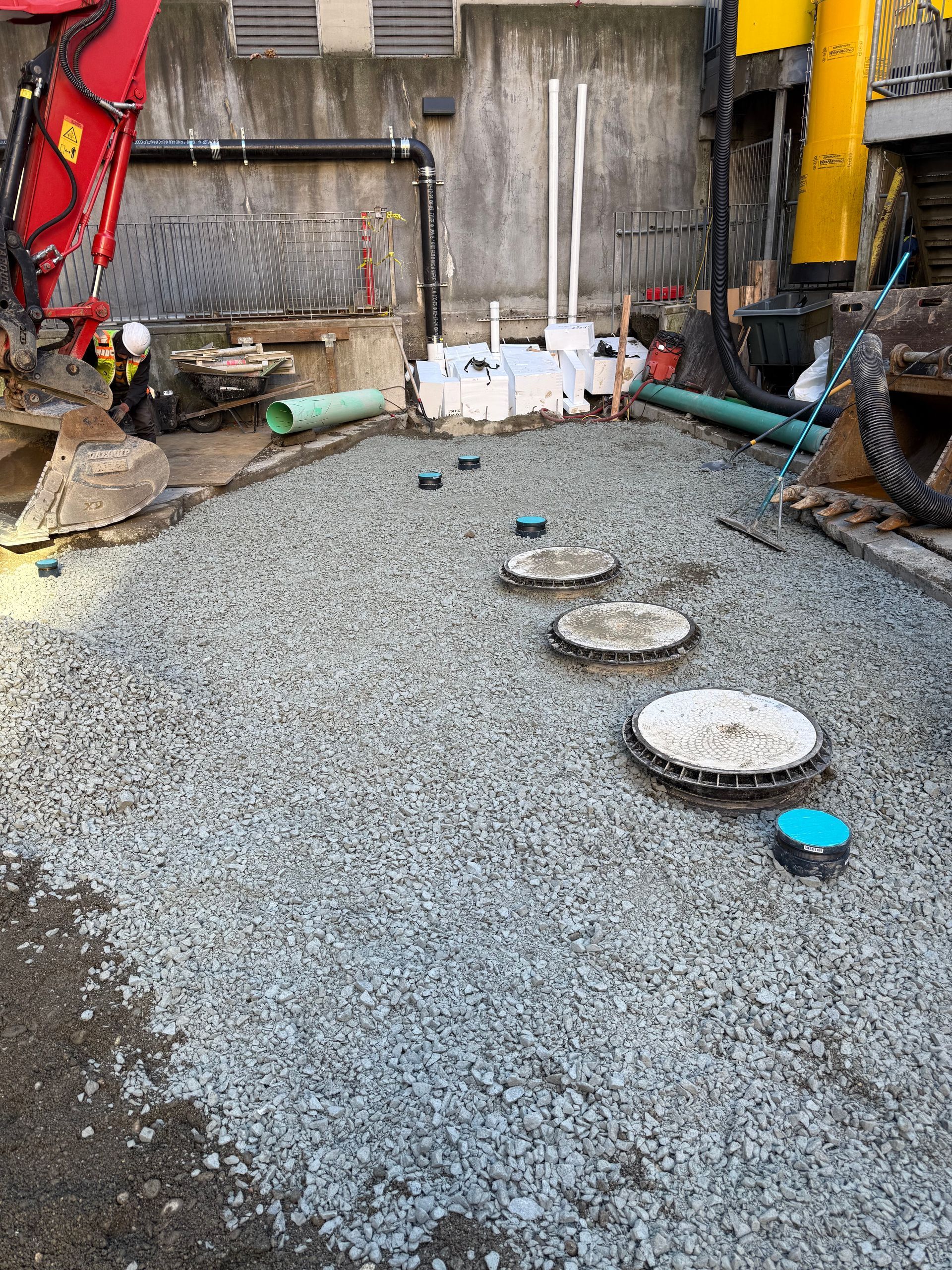 A gravel-covered construction site with several circular maintenance access covers and pipes protruding from the ground.
