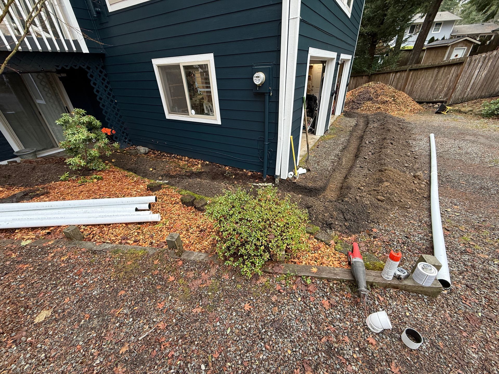 A navy-blue house exterior with a trench dug along the side, white PVC pipes, and construction tools on a gravel yard.