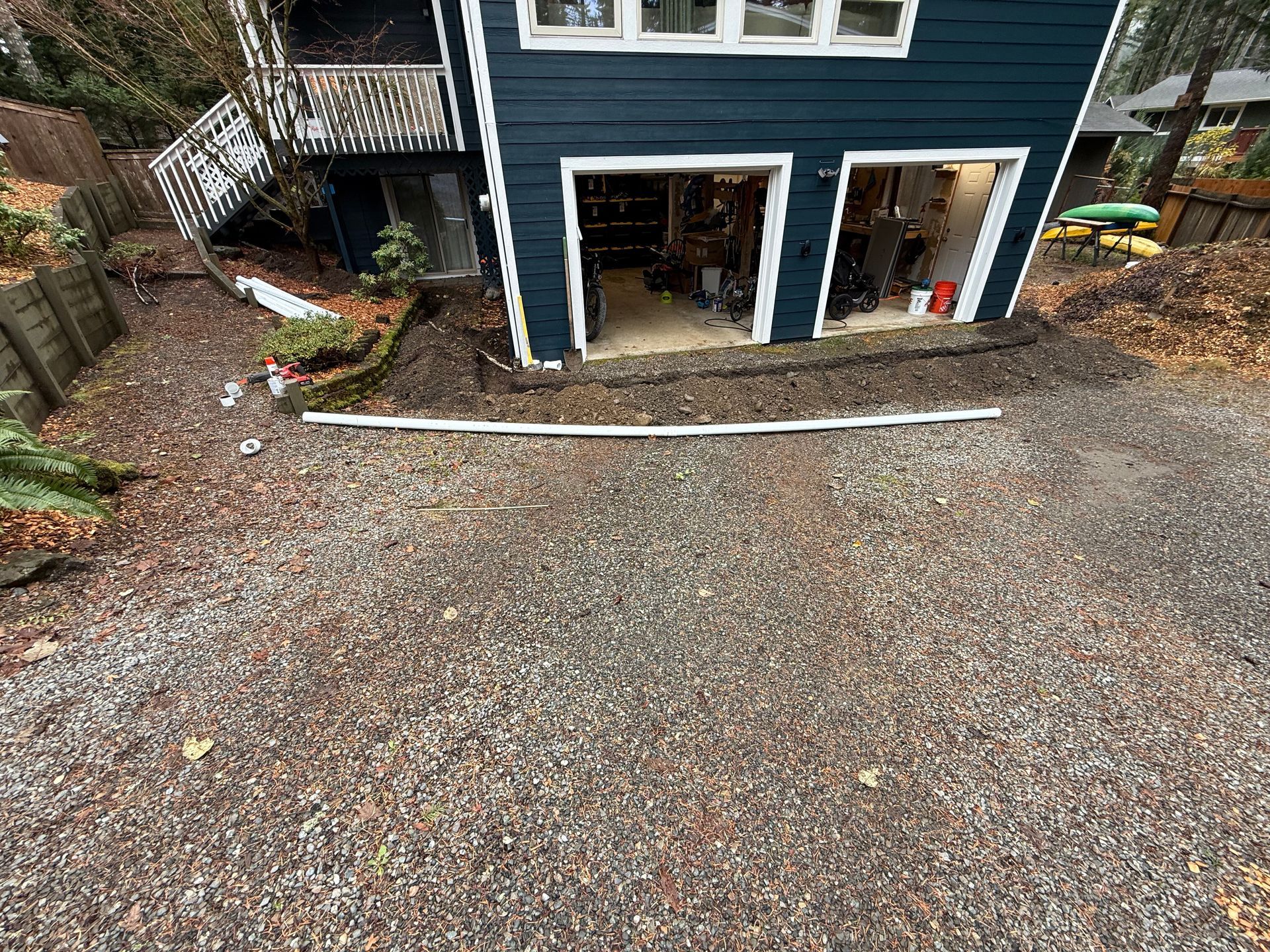 A blue house with an open two-car garage sits behind a gravel driveway with a white pipe lying across the ground.