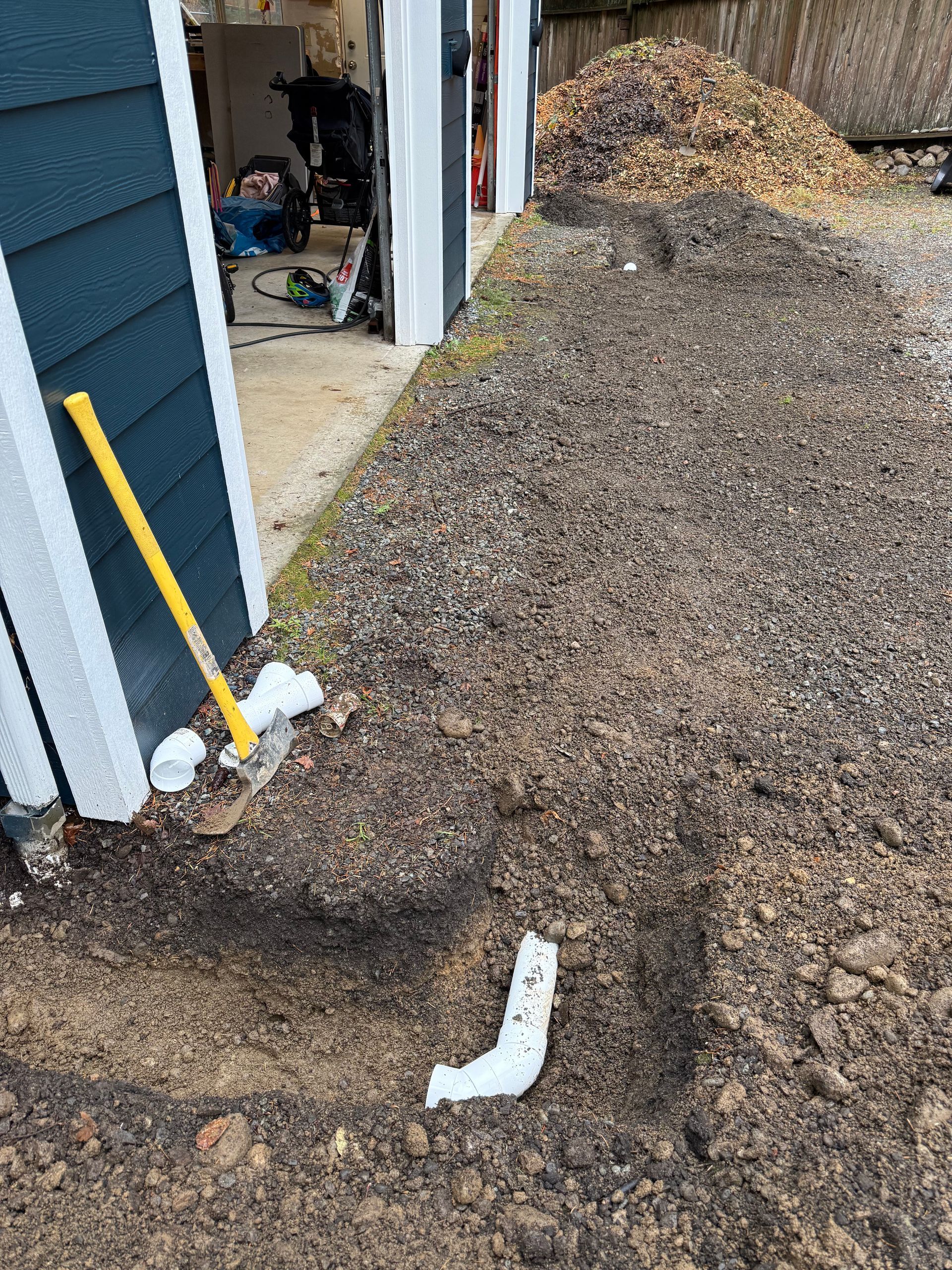 A trench with white PVC pipe sections near the exterior wall of a garage under construction, with soil piles nearby.