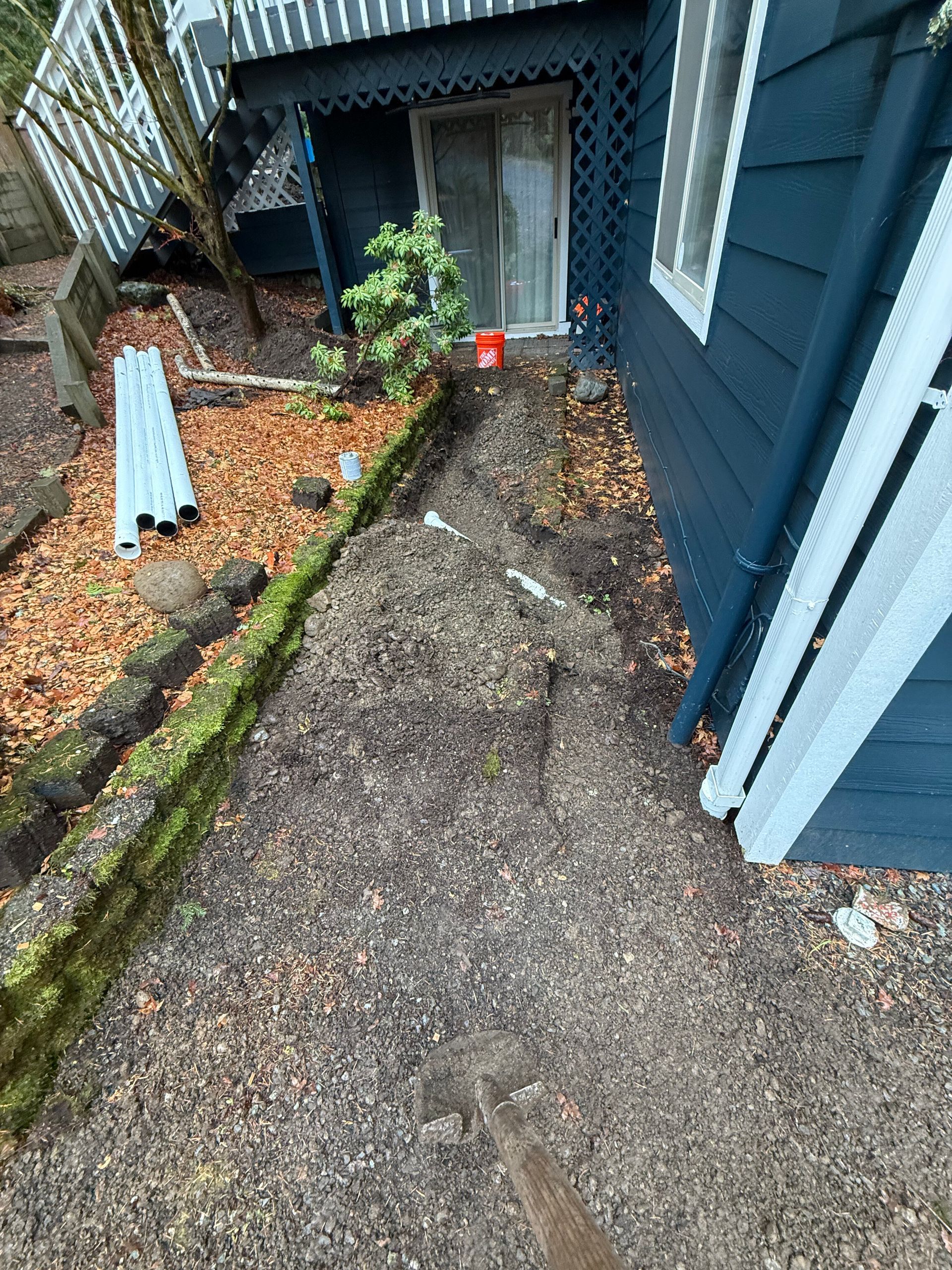 A construction area with gravel, a pipe trench, and building materials next to the blue siding of a house.