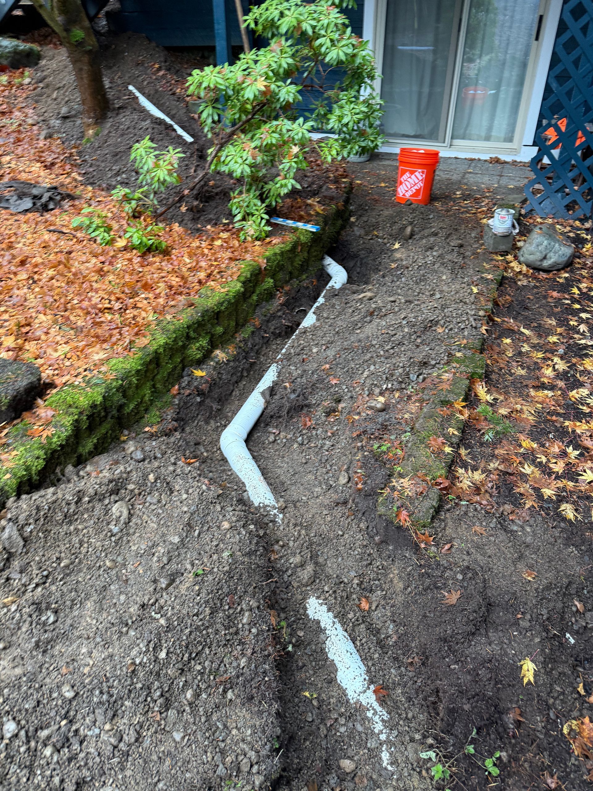 A trench with white drainage pipe segments and a red bucket near a patio door in a garden setting.