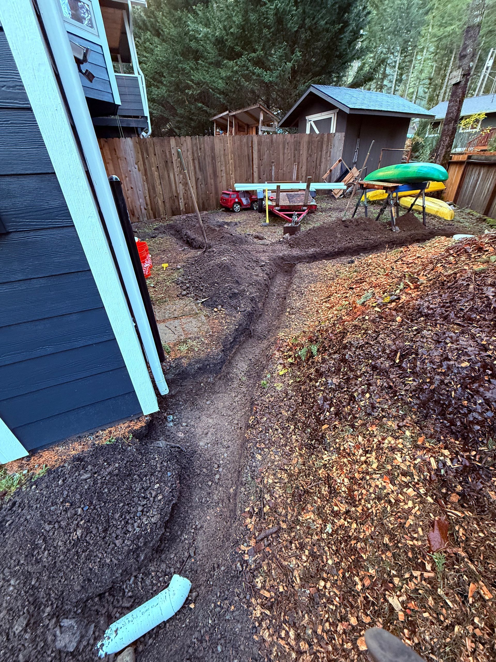 A trench dug in a residential yard beside a dark blue house, leading toward a shed and yard equipment in the background.