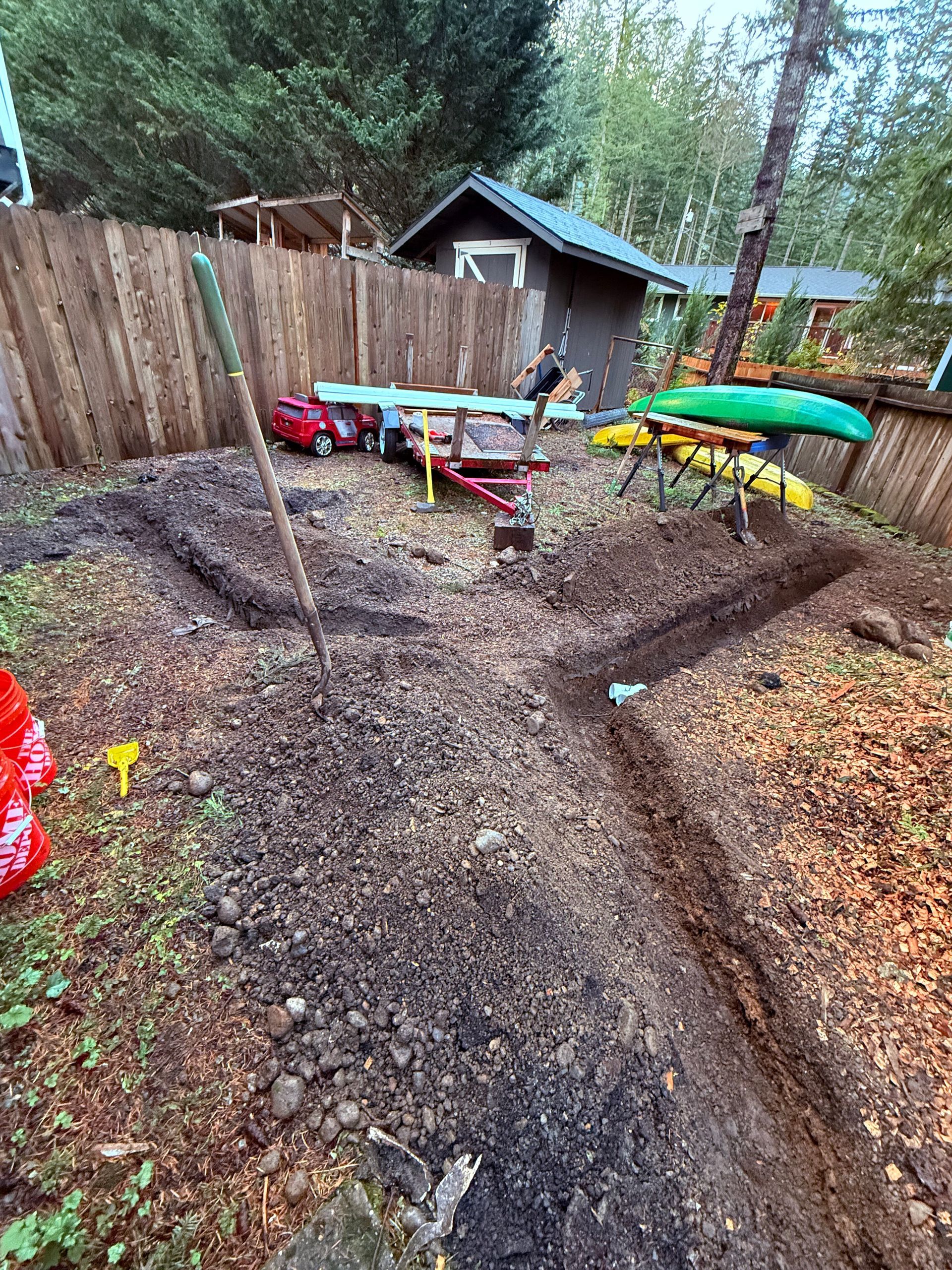 A dirt yard with two trenches dug into the ground, a shovel, a small trailer, and canoes on racks near a wooden shed.