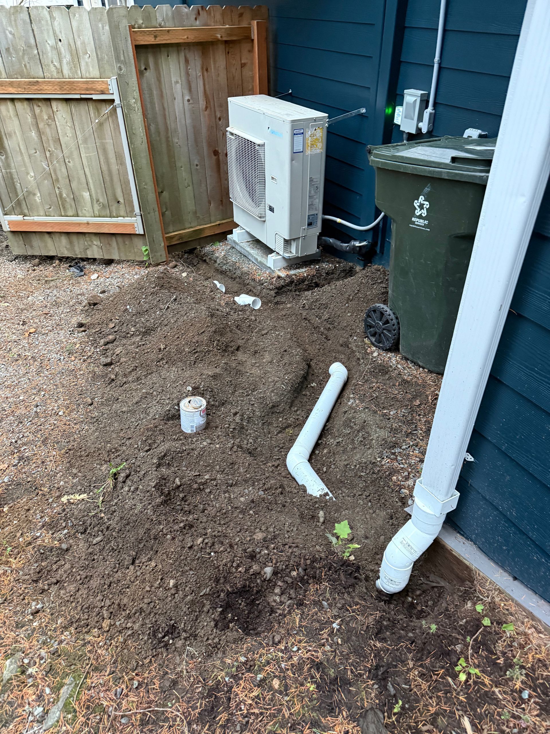 A backyard area with a heat pump unit, a green trash bin, and two exposed white pipes protruding from the soil.