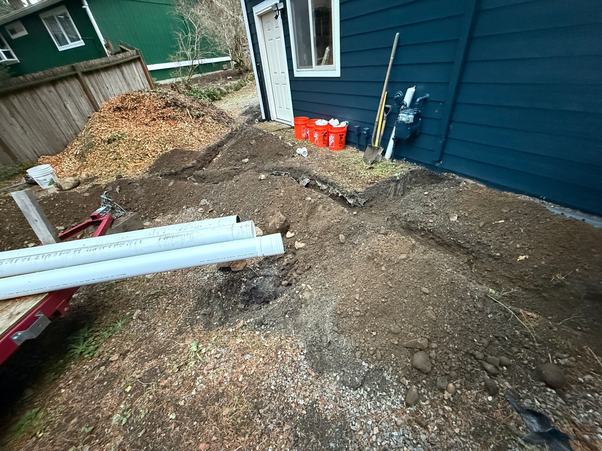 A trench is being dug in a residential yard next to a dark blue house, with PVC pipes on a trailer nearby.