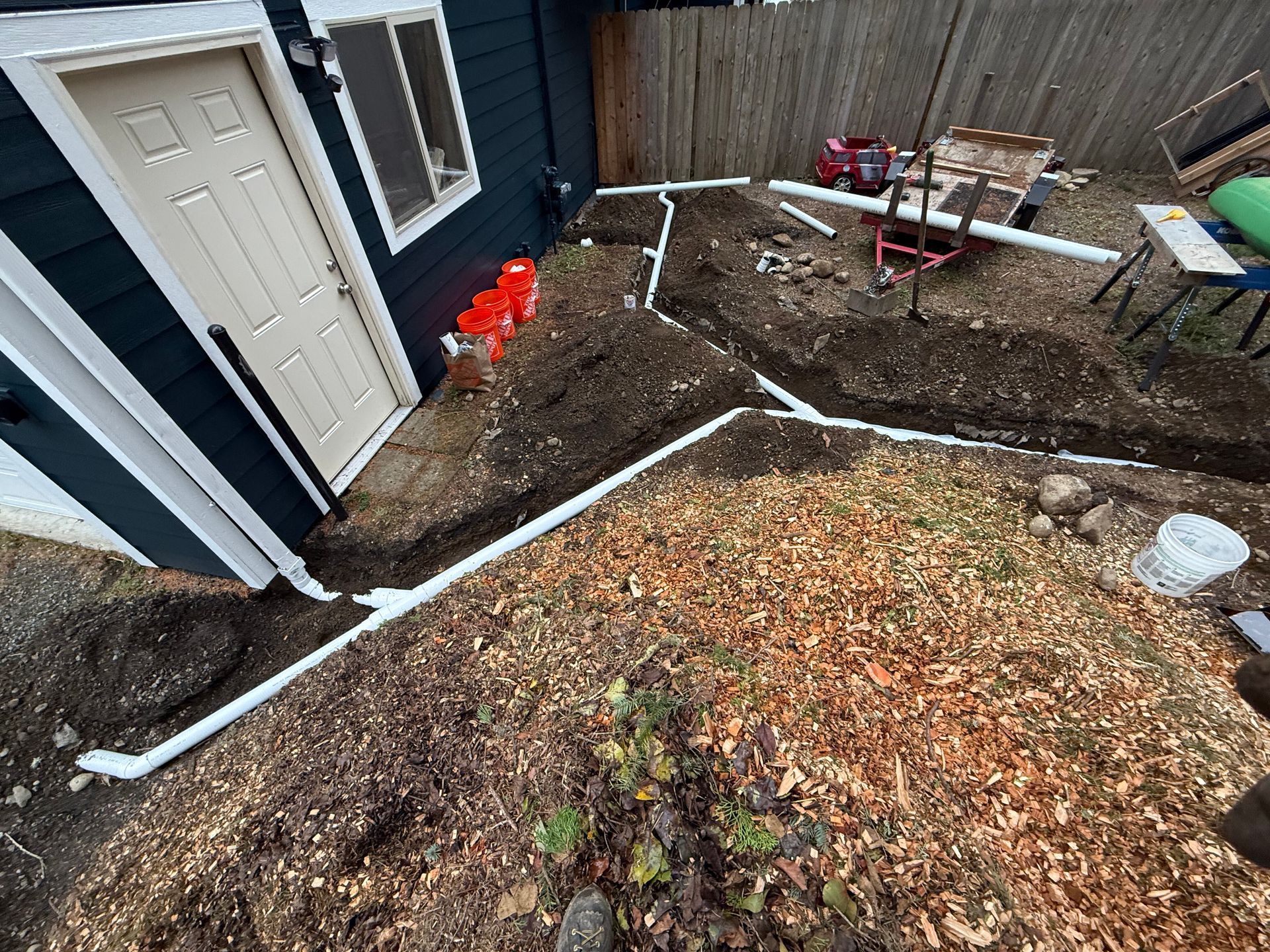 A backyard under renovation with white PVC drainage pipes laid in trenches along a dark blue house and wood-chipped yard.