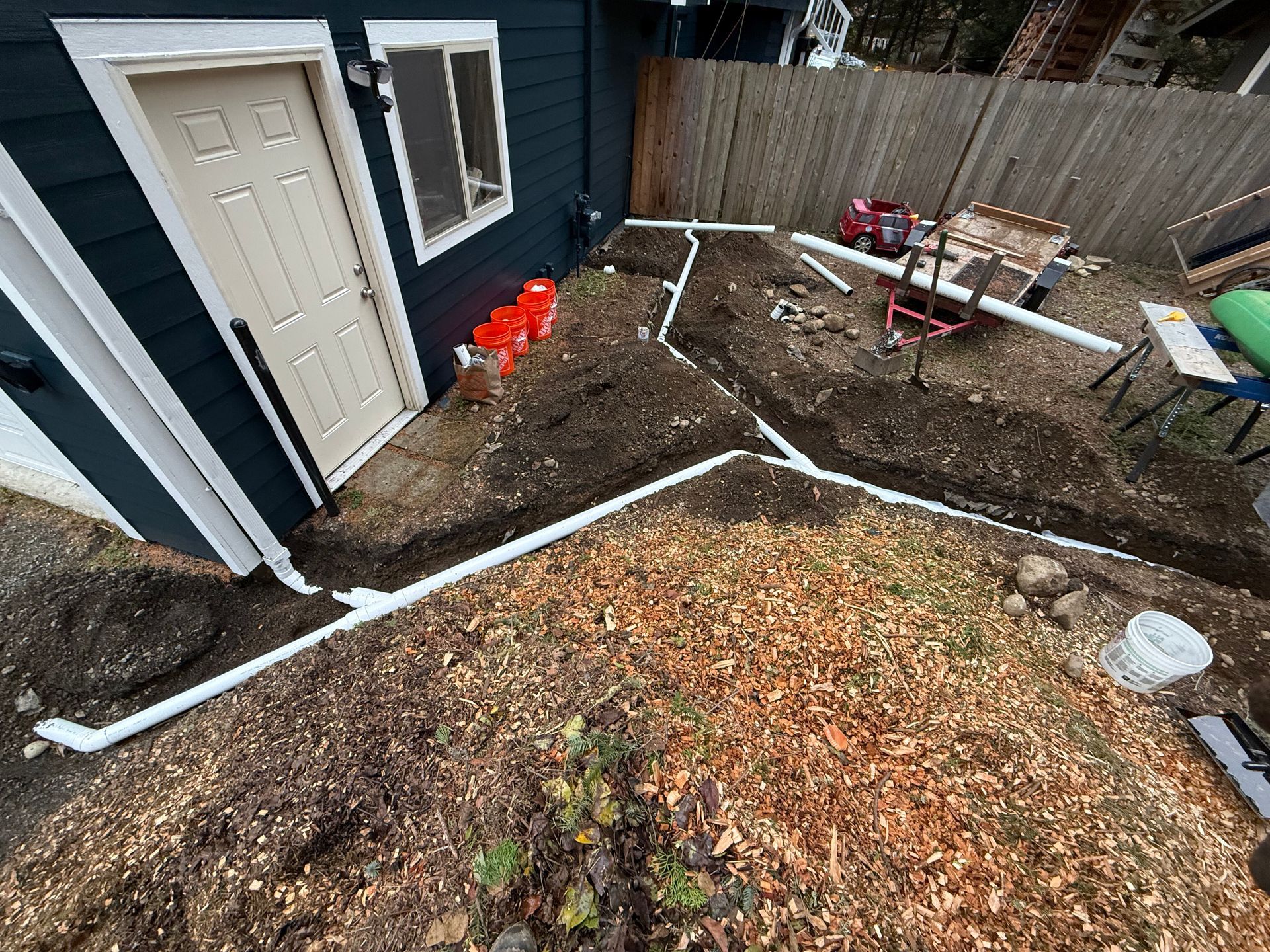 White PVC drainage pipes laid in trenches across a yard next to a dark blue house with a beige door.