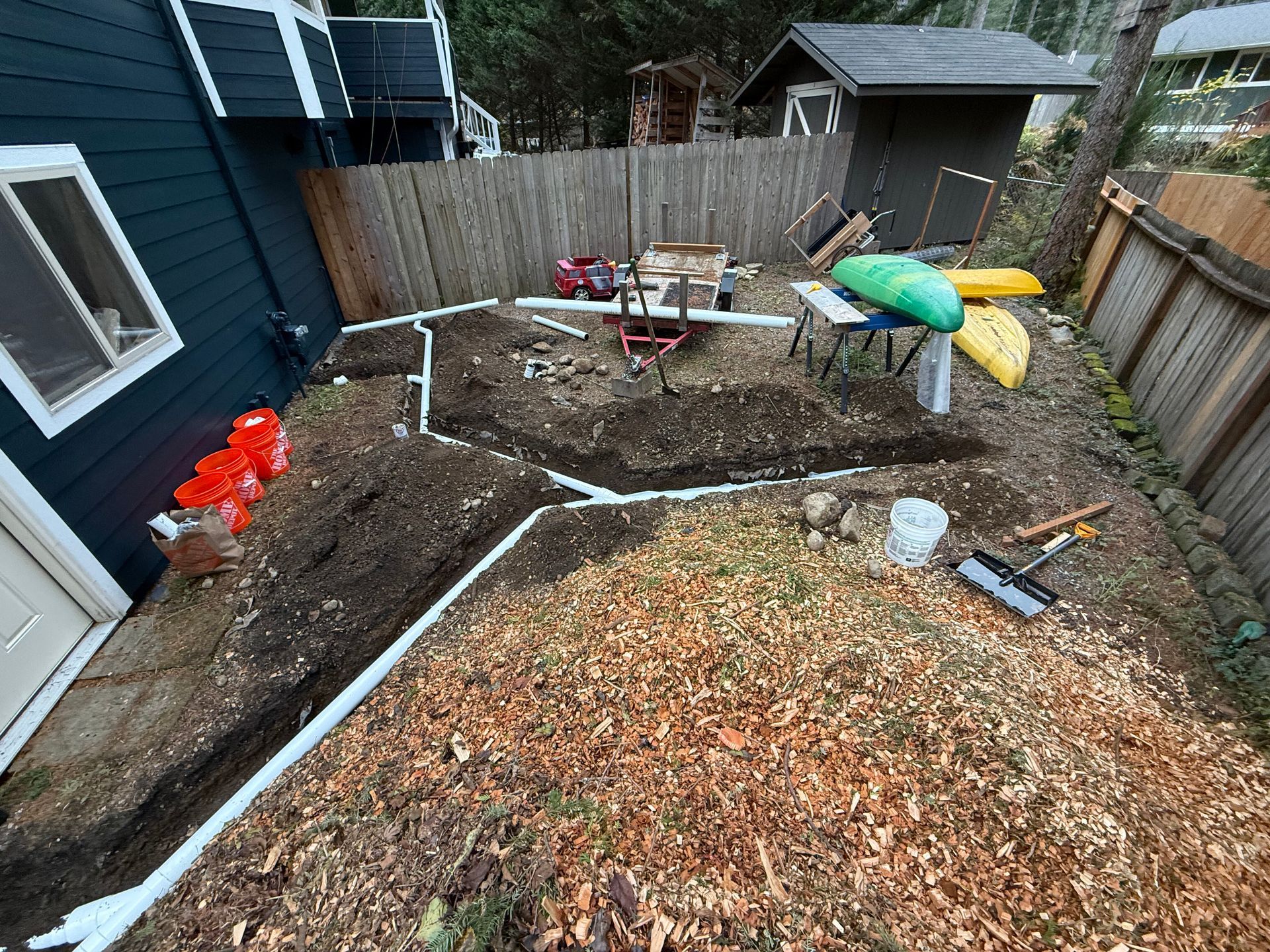 A backyard renovation project featuring newly installed white PVC drainage pipes in trenches near a dark blue house.