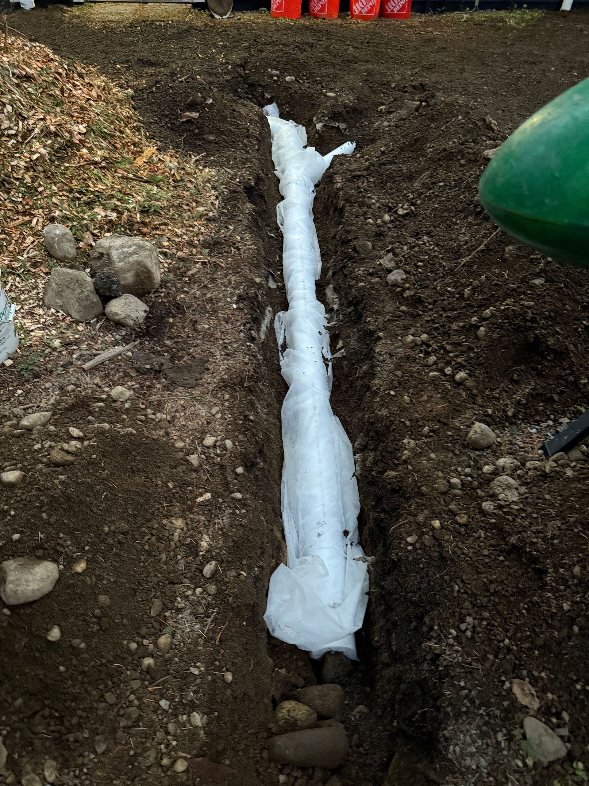 A white, fabric-wrapped drainage pipe lies centered in a narrow, freshly dug dirt trench outdoors.