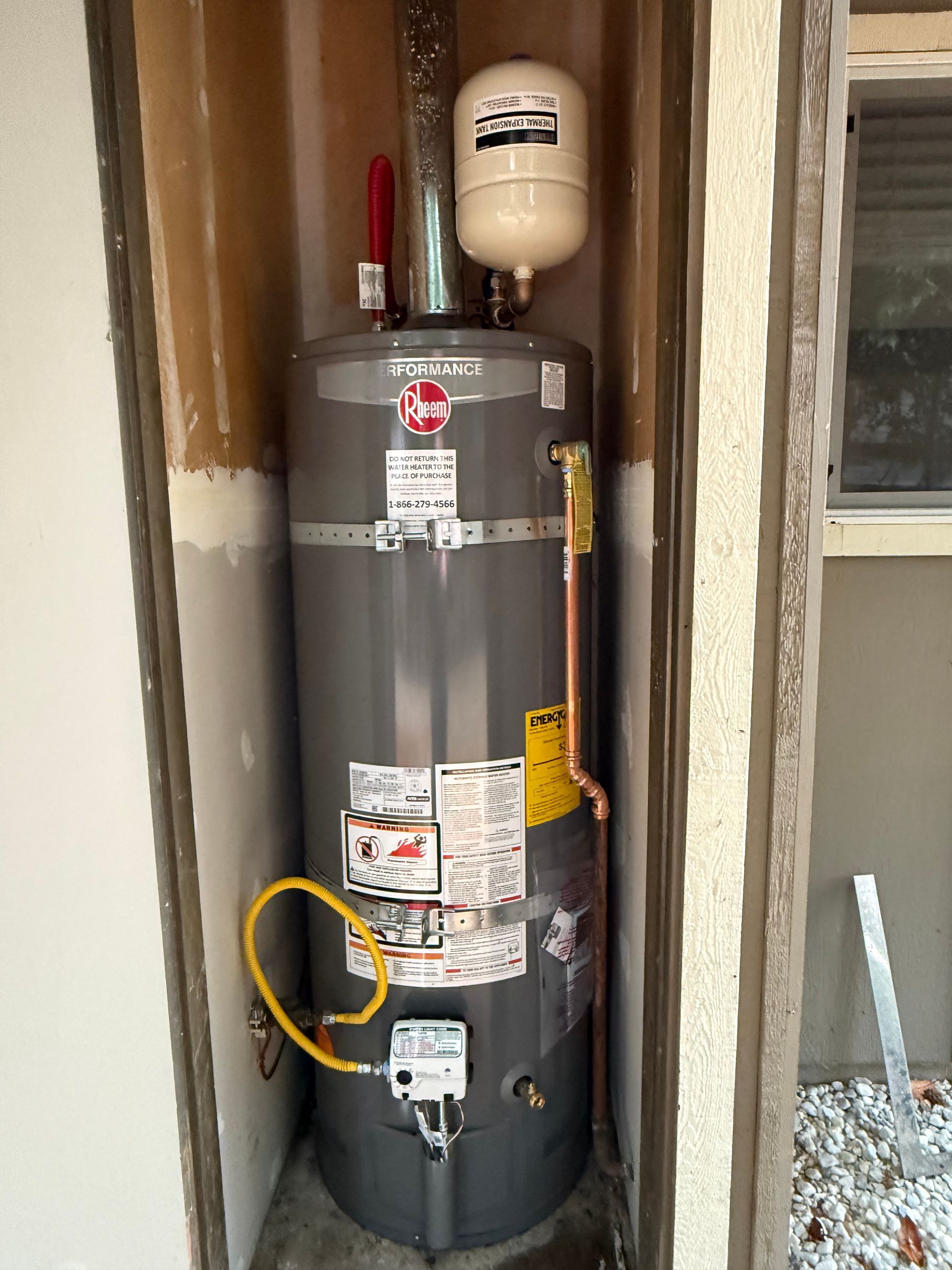 A grey Rheem gas water heater installed in an outdoor closet, featuring an attached expansion tank and yellow gas line.