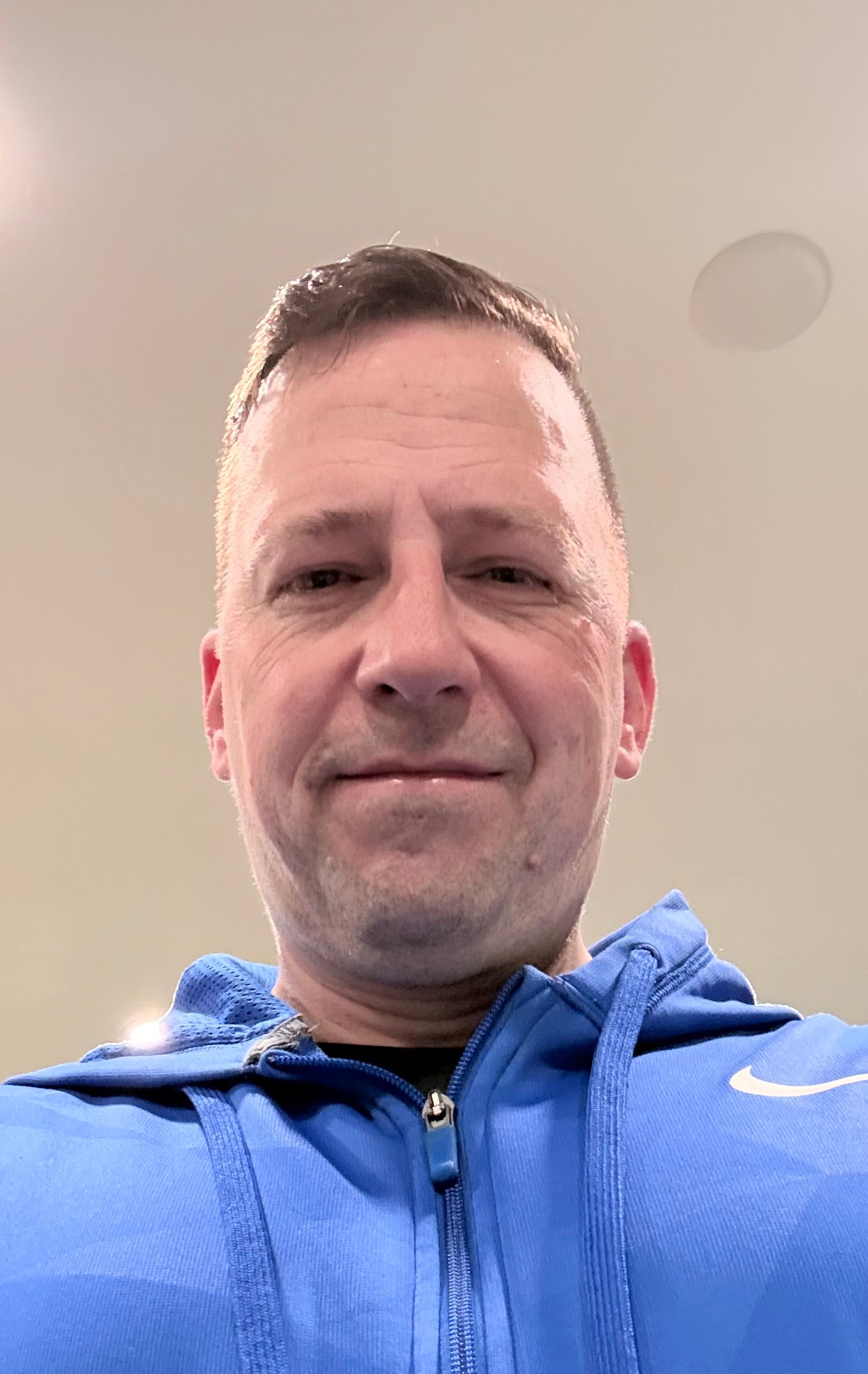 A person with short hair wearing a blue zip-up athletic hoodie, looking directly at the camera.