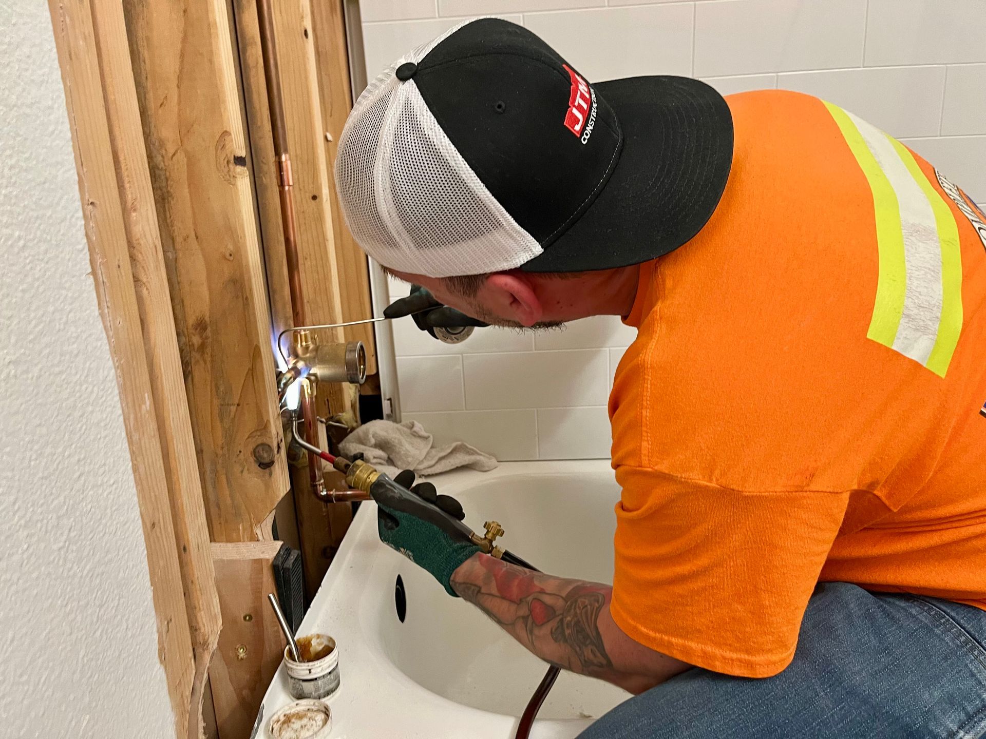 A plumber in an orange shirt uses a blowtorch to solder copper pipes in a wall opening above a white bathtub.