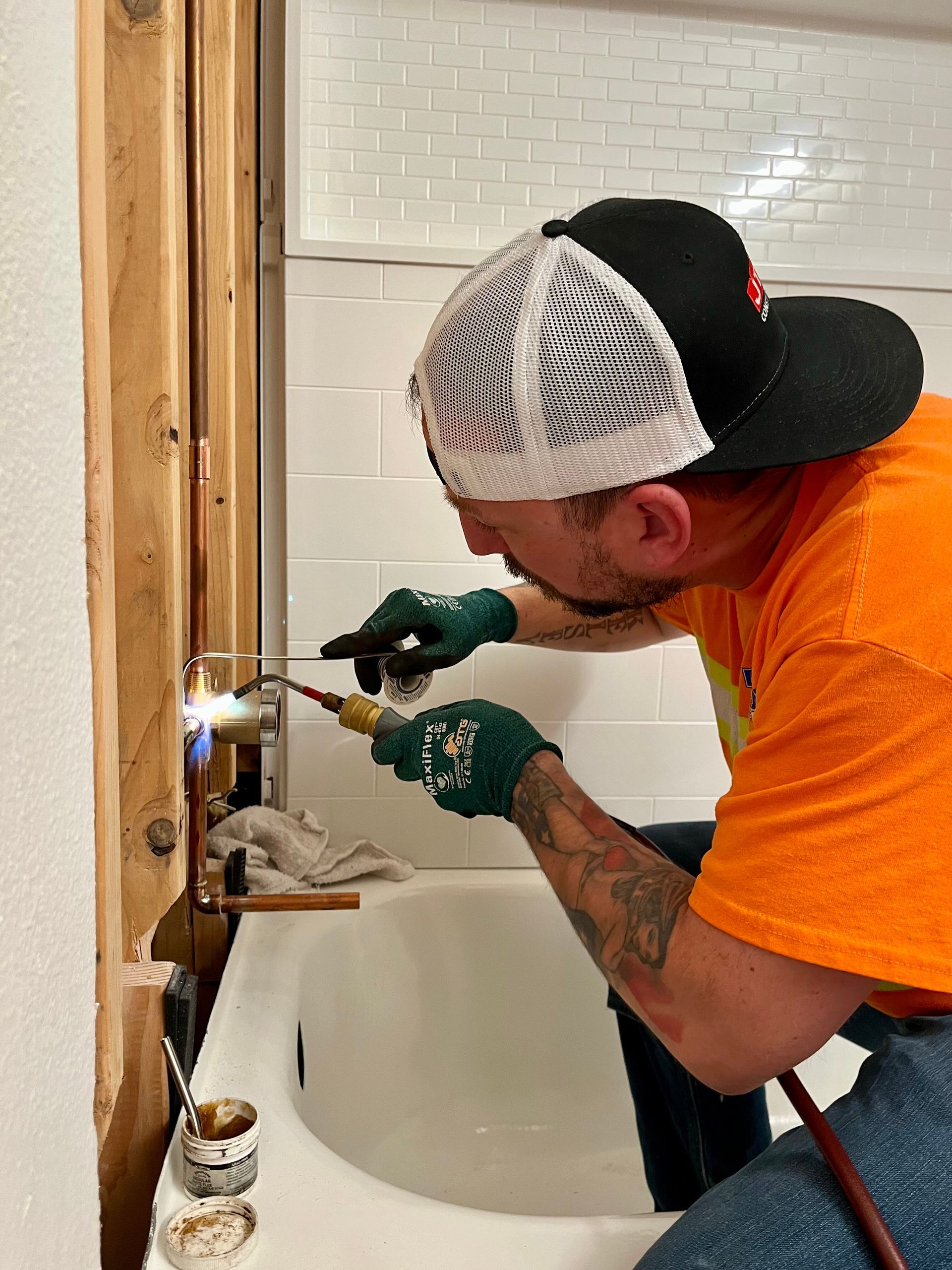 A person in an orange shirt and green gloves uses a torch to solder copper pipes near a bathtub in a tiled alcove.