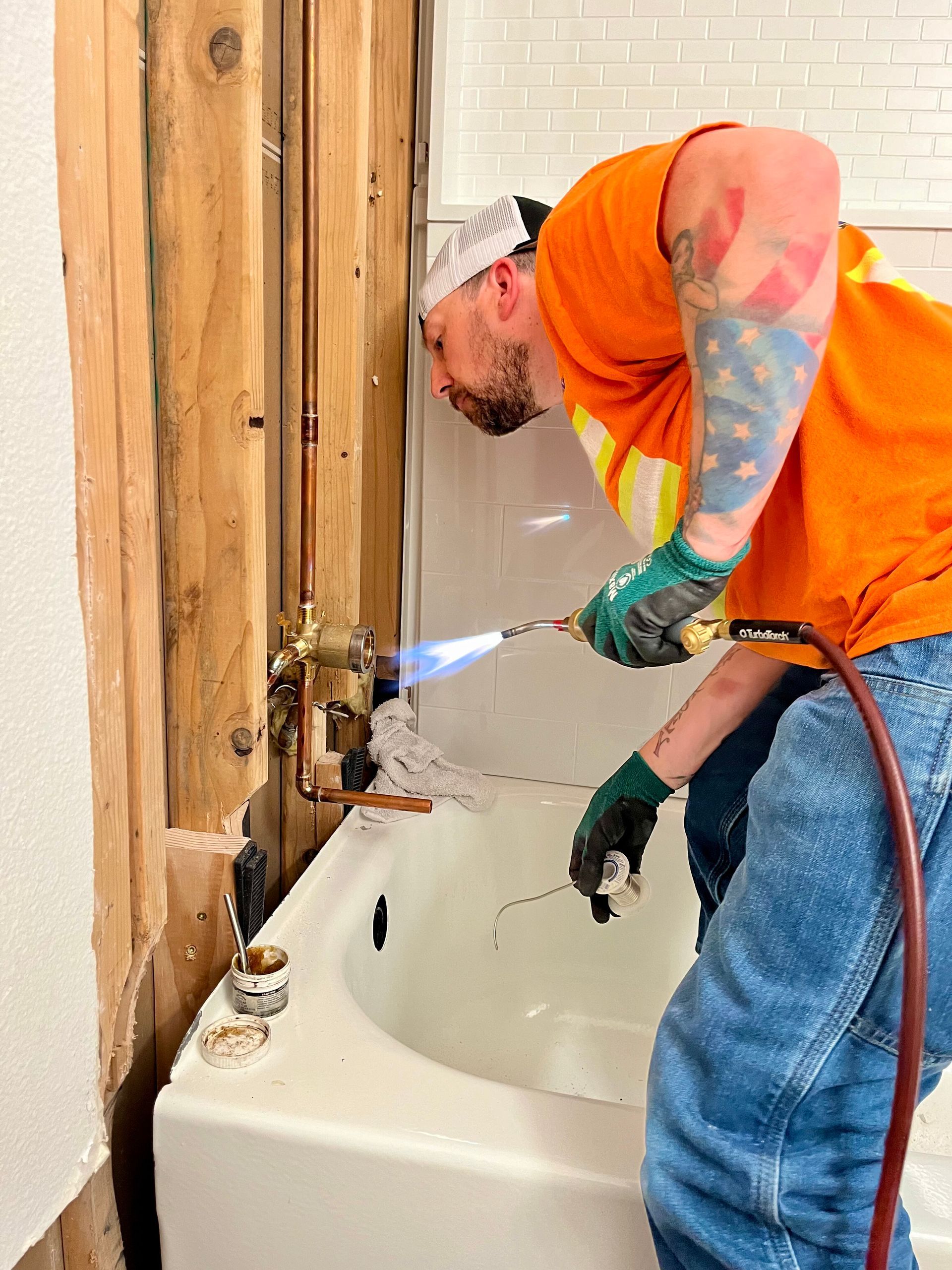 A person wearing an orange shirt and gloves uses a torch to solder copper pipes inside an open wall above a bathtub.