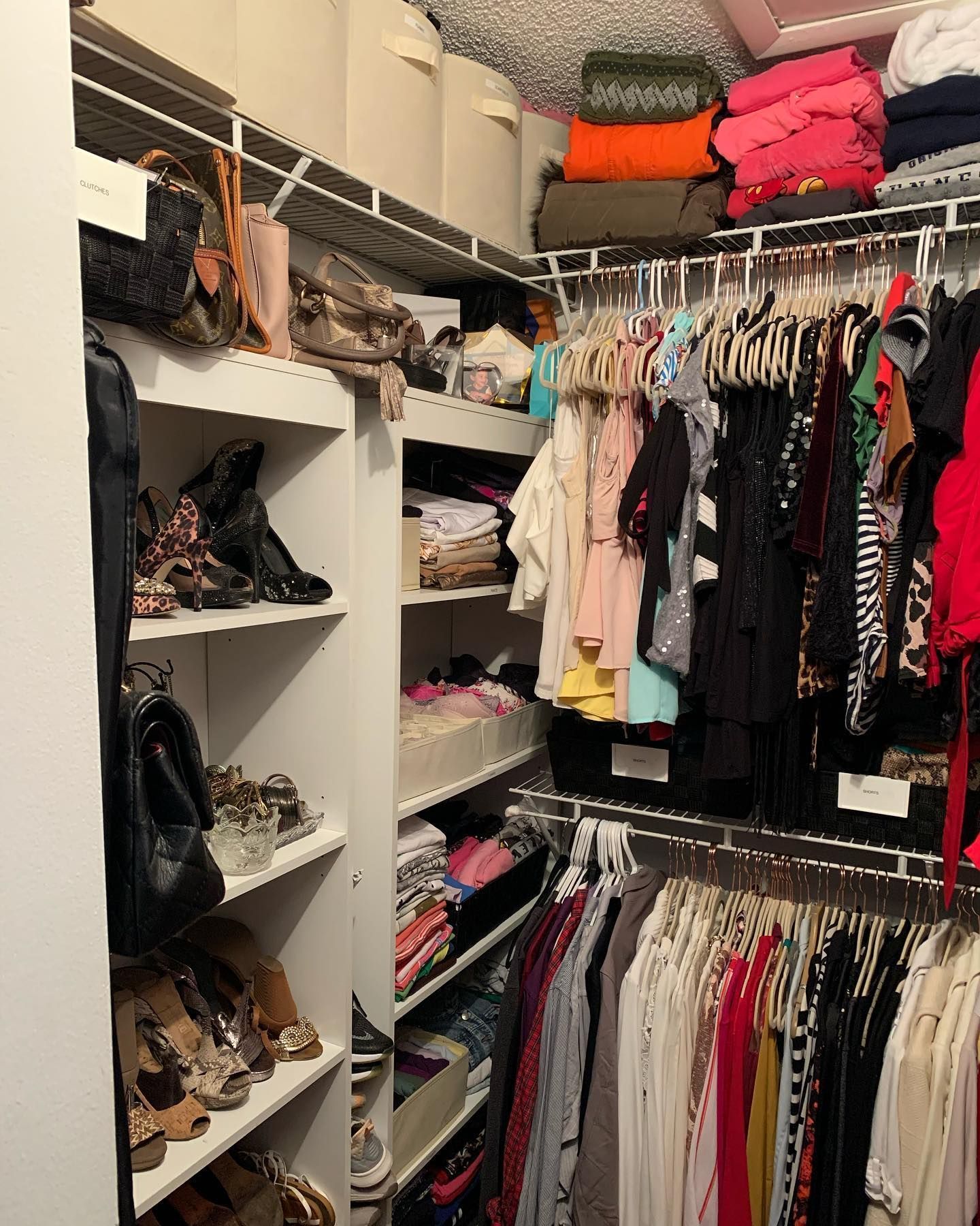Walk-in closet with clothes on hangers and stacked shelves of shoes and folded items.