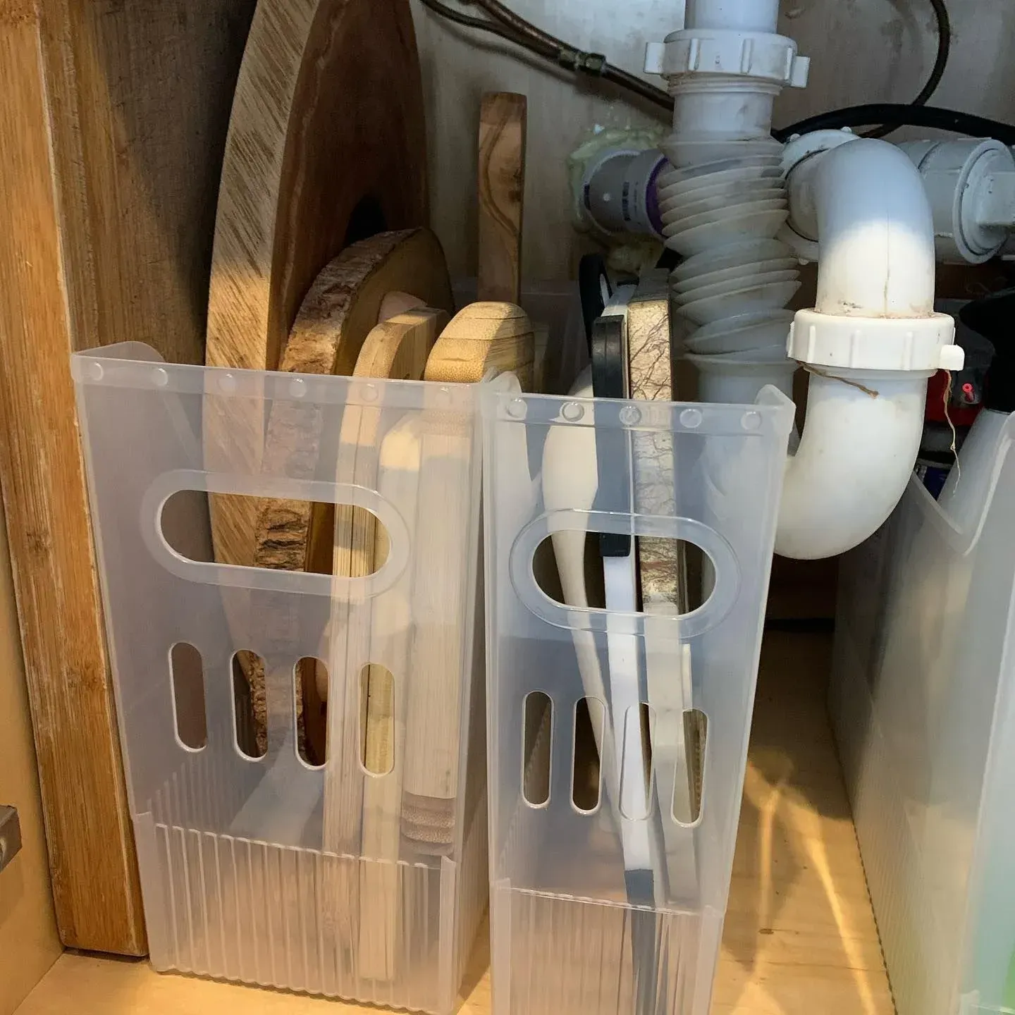 Two clear plastic bins filled with wooden cutting boards and knives under a sink.